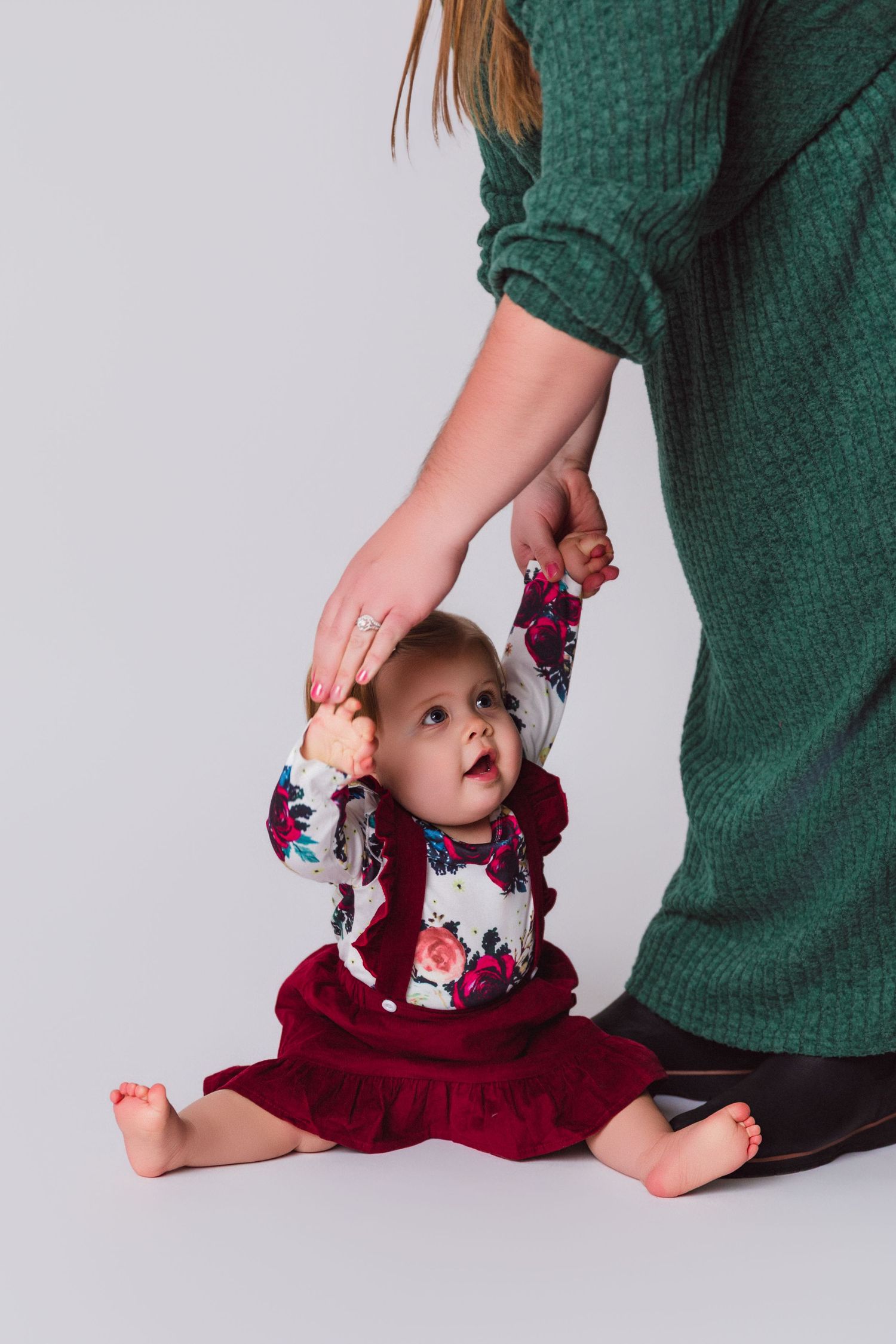 Baby wearing burgundy outfit reaches up while being supported by someone in green.