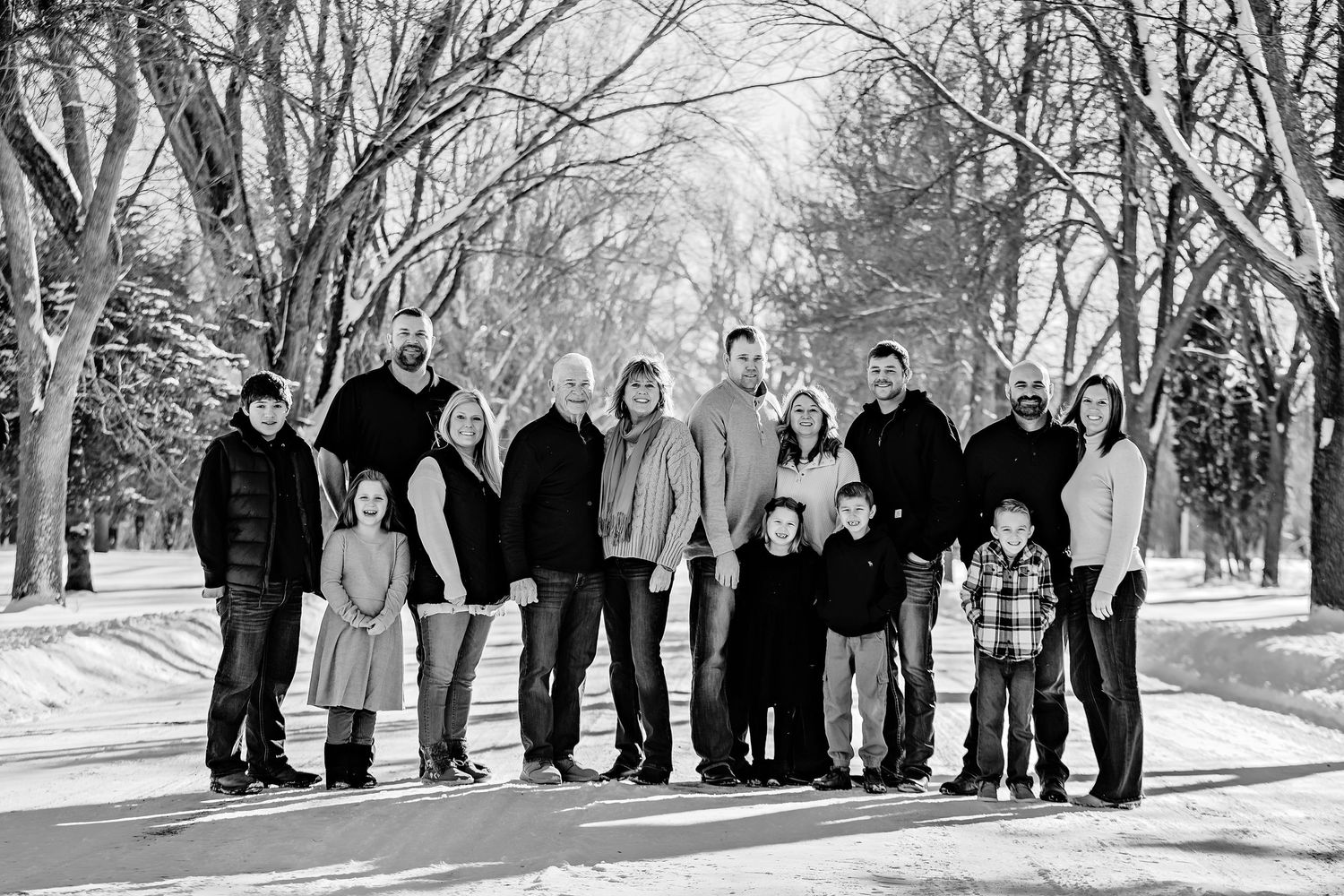 Extended family gathered for a winter portrait on a snow-covered walkway surrounded by bare trees.