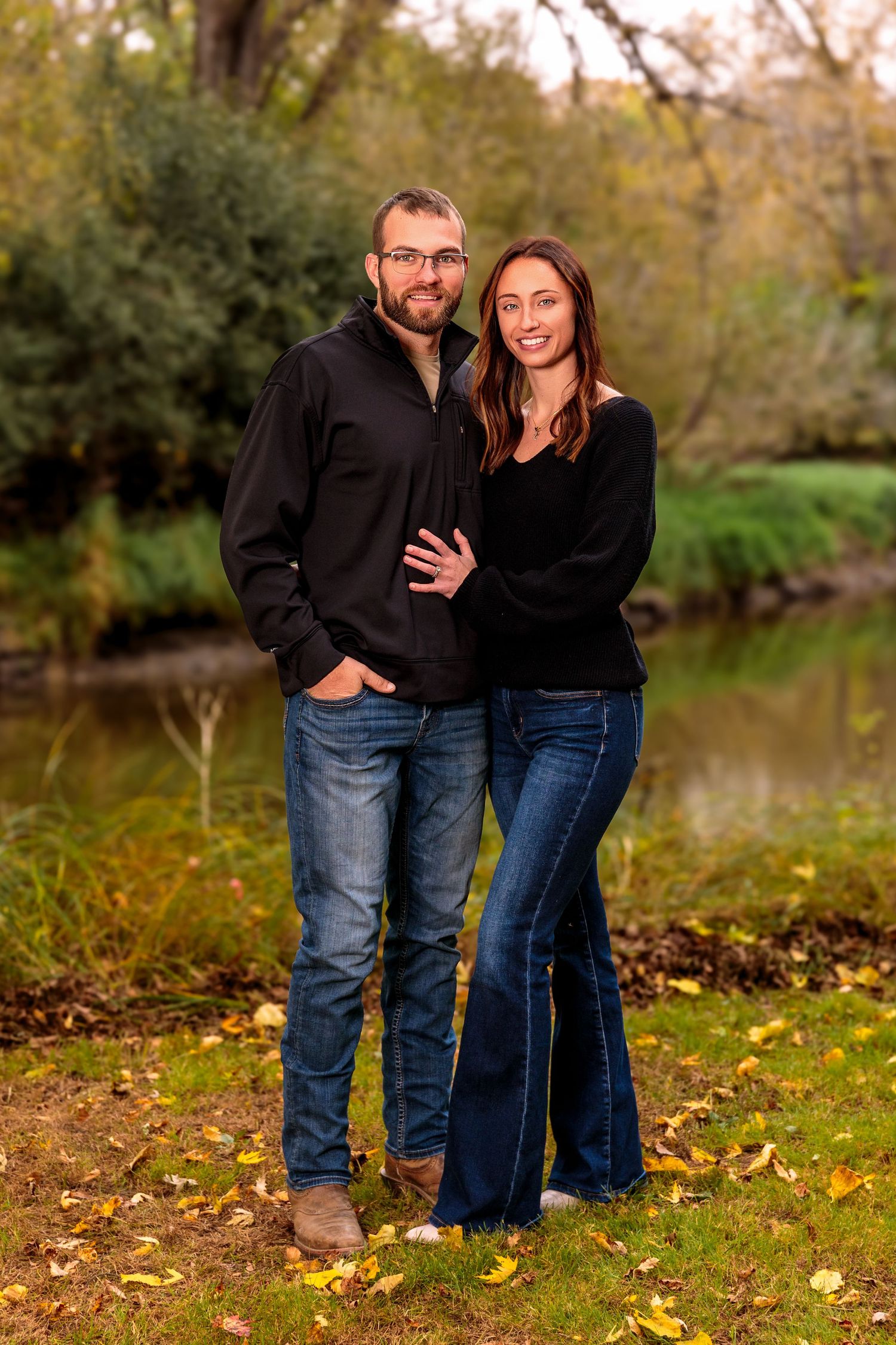 Couple in casual wear embracing while standing on fallen autumn leaves near a pond.
