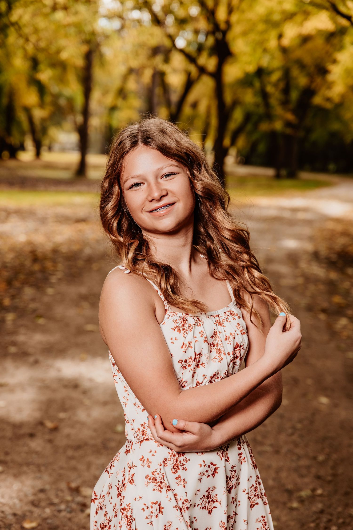 A person in a floral sundress poses outdoors in a park with autumn trees in the background.