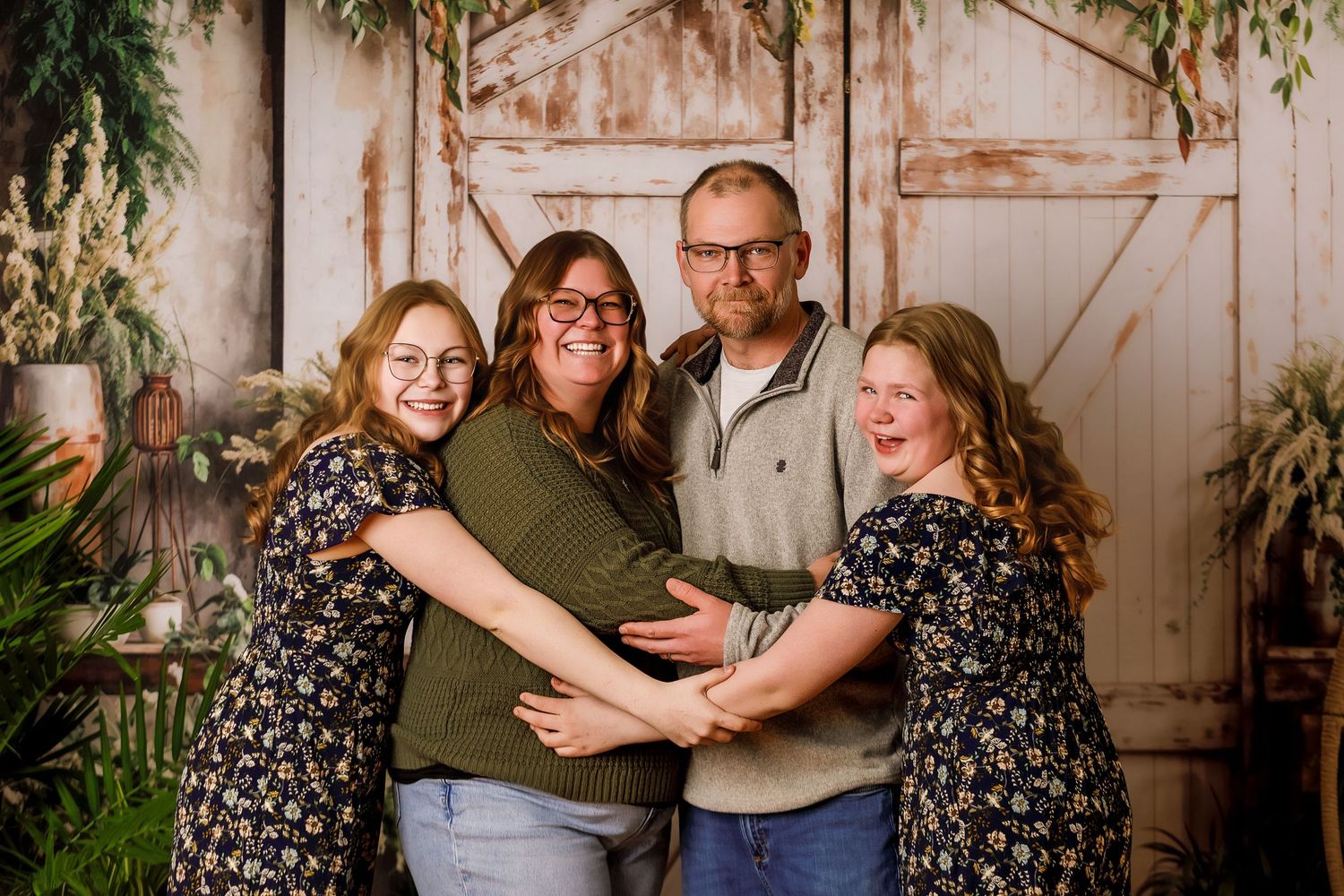 Family of four posing and smiling together in front of rustic barn doors with greenery backdrop.