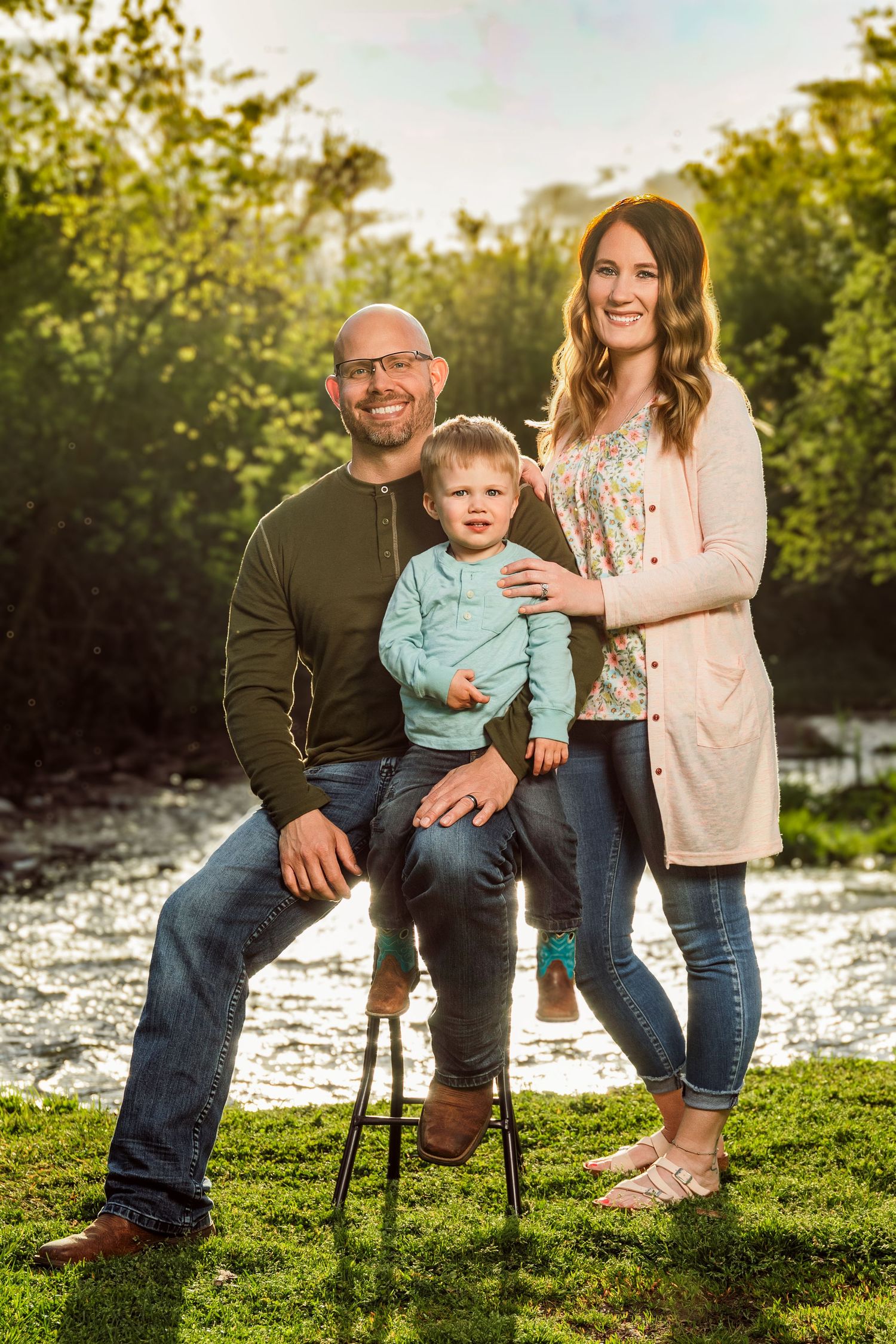 Family portrait of parents with toddler outdoors by a stream, surrounded by lush green trees in warm golden light.