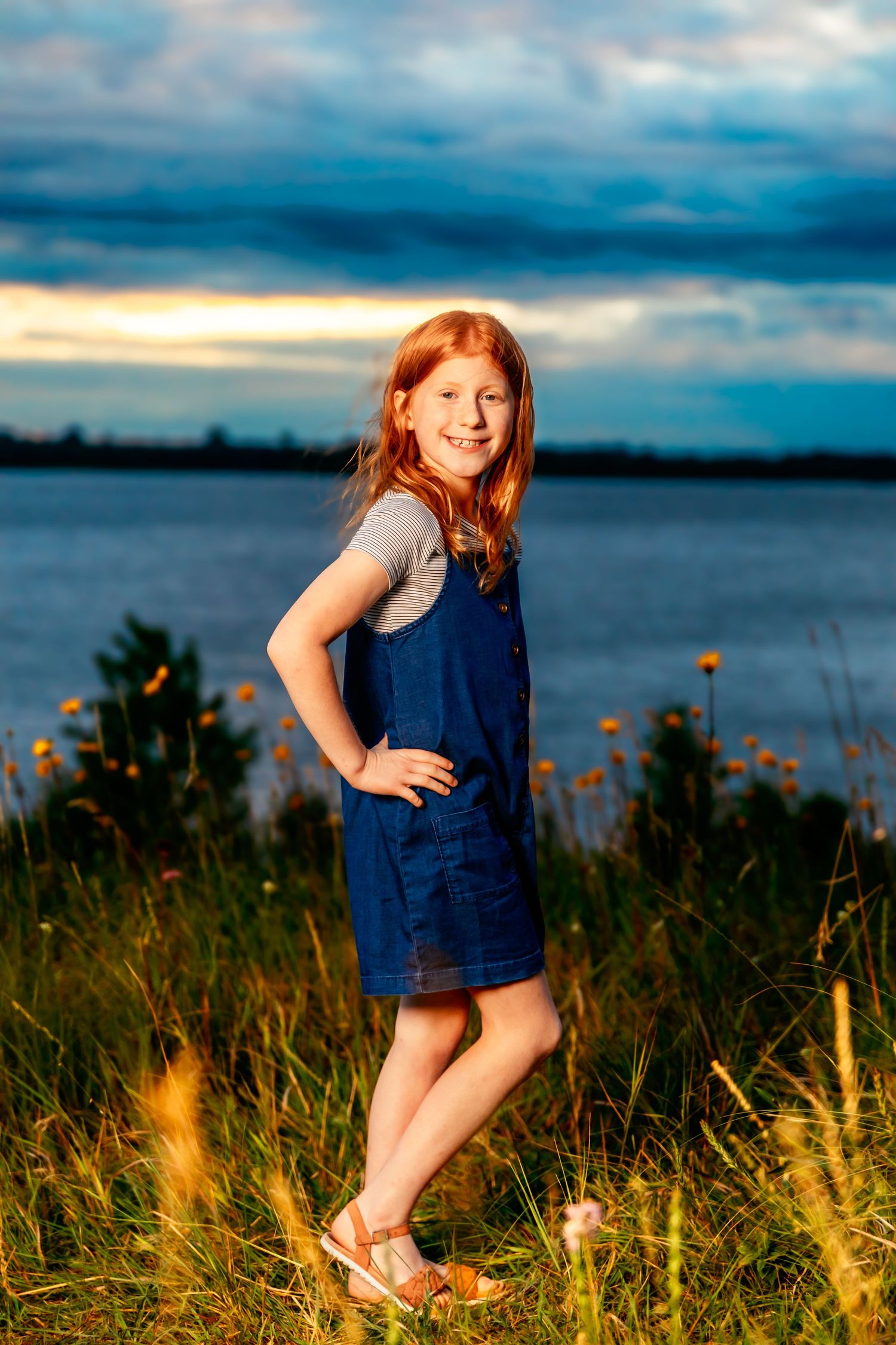 A joyful figure in a blue dress stands near wildflowers by a lake at sunset.