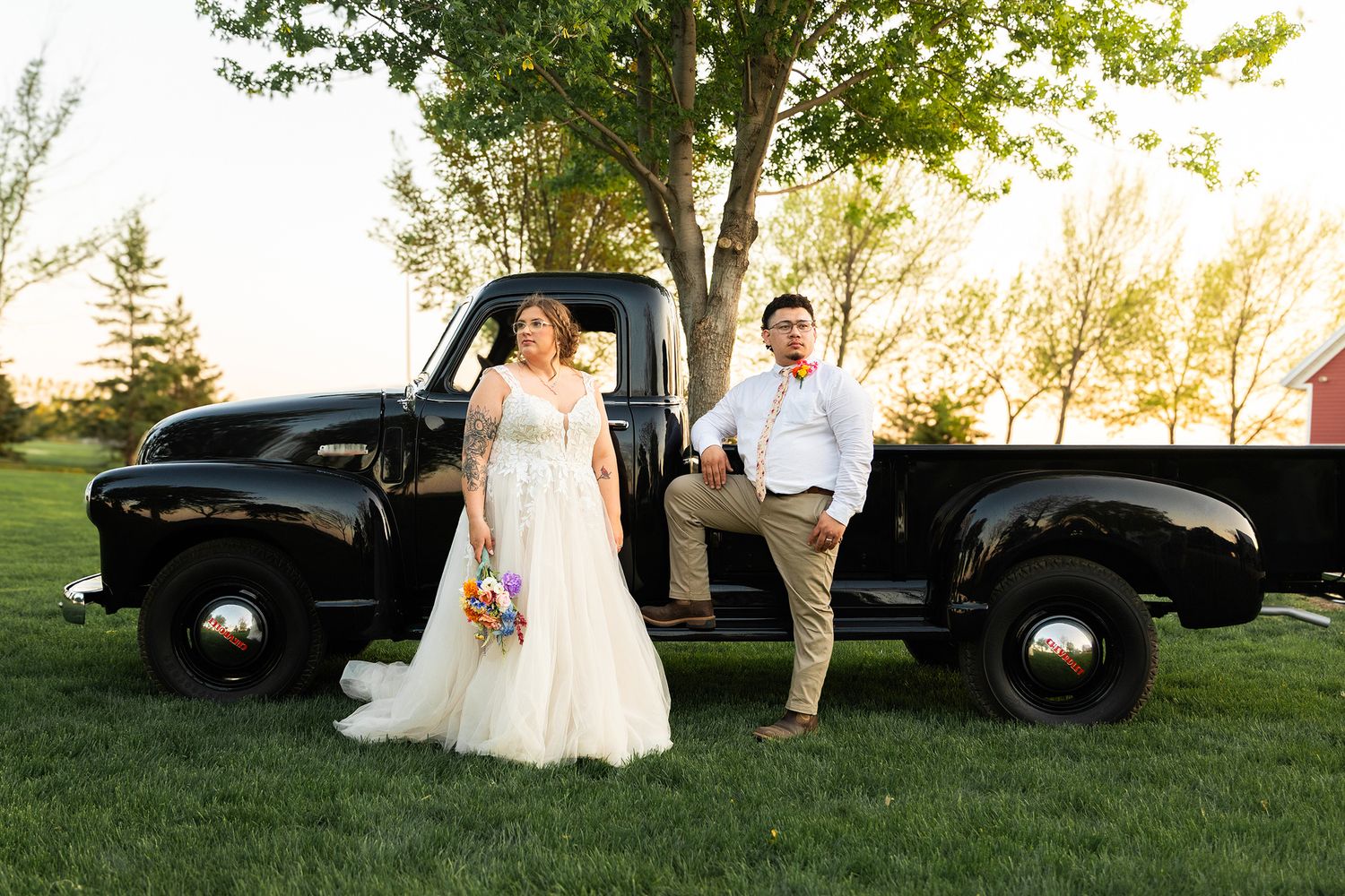 A wedding couple poses with a vintage black pickup truck on a sunlit grassy lawn.