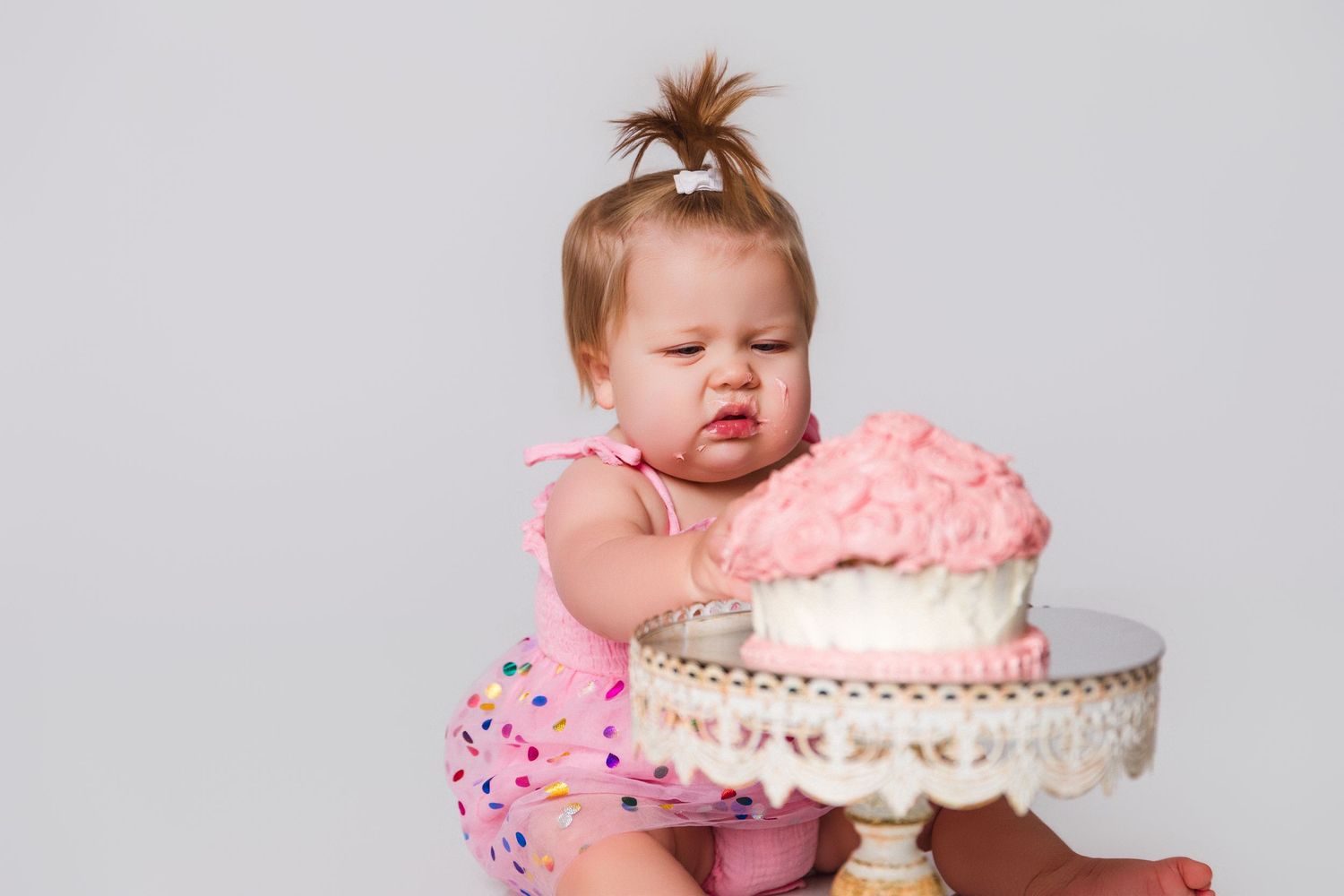 A small child in a pink dress reaches for a decorated cake on a pedestal.