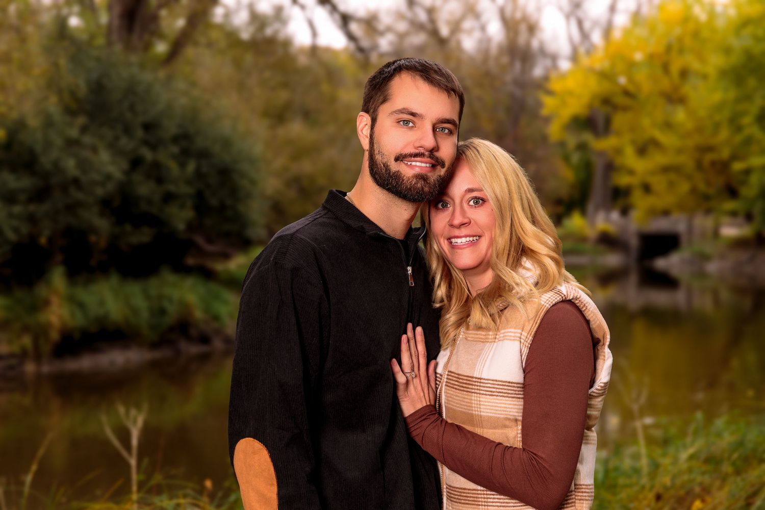 Couple embracing outdoors near lake with autumn trees in background.