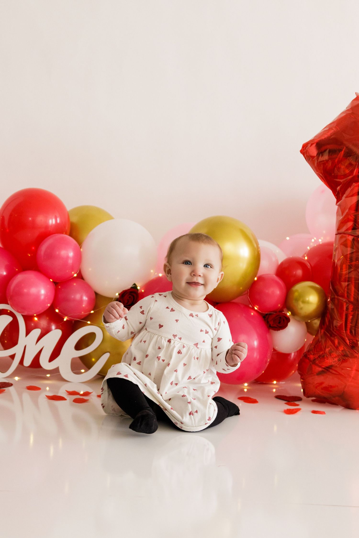 Festive Valentine's day setup with red and gold balloons, white decorations and a small figure in a floral outfit.