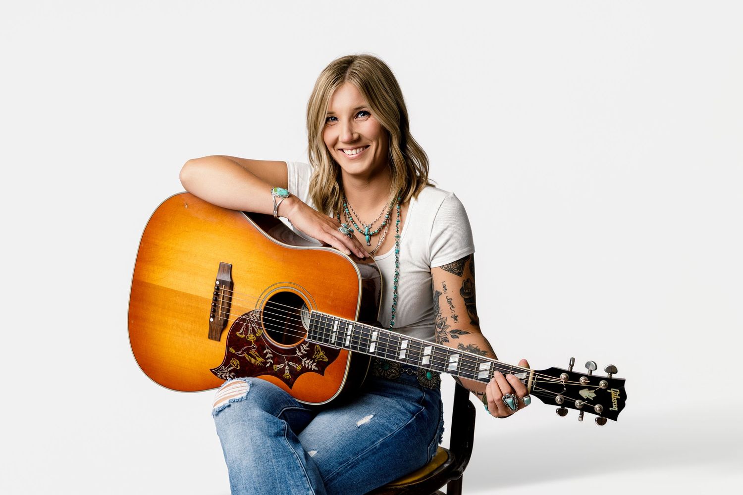 Smiling female guitarist with tattoos holding acoustic guitar, wearing white tee and jeans, seated against white background.