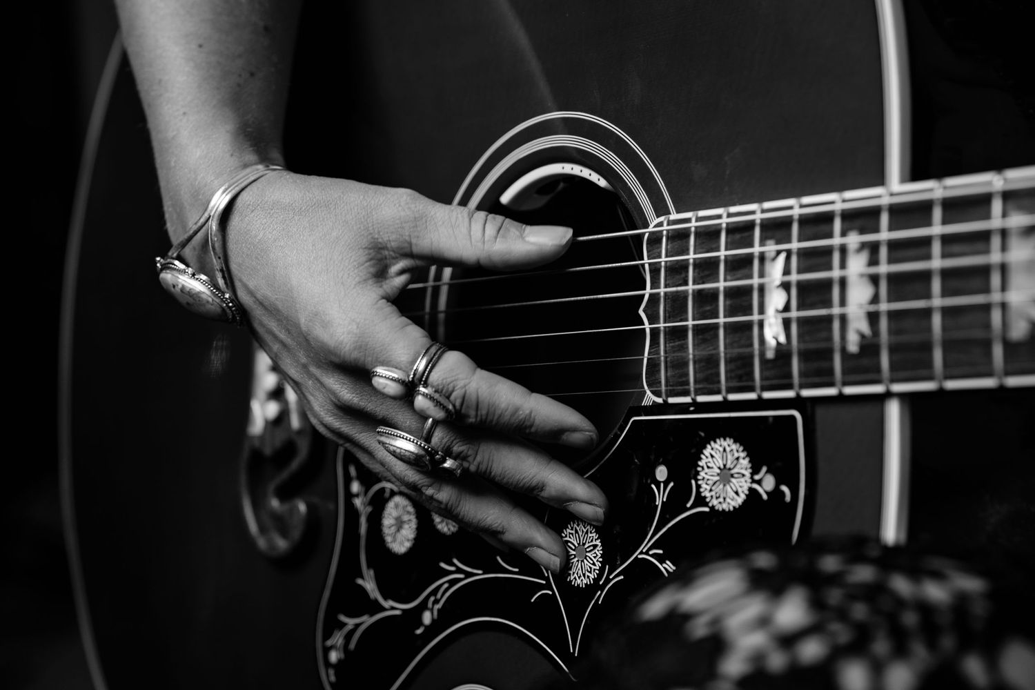 Black and white close-up of a woman's hand with rings strumming a decorated acoustic guitar.