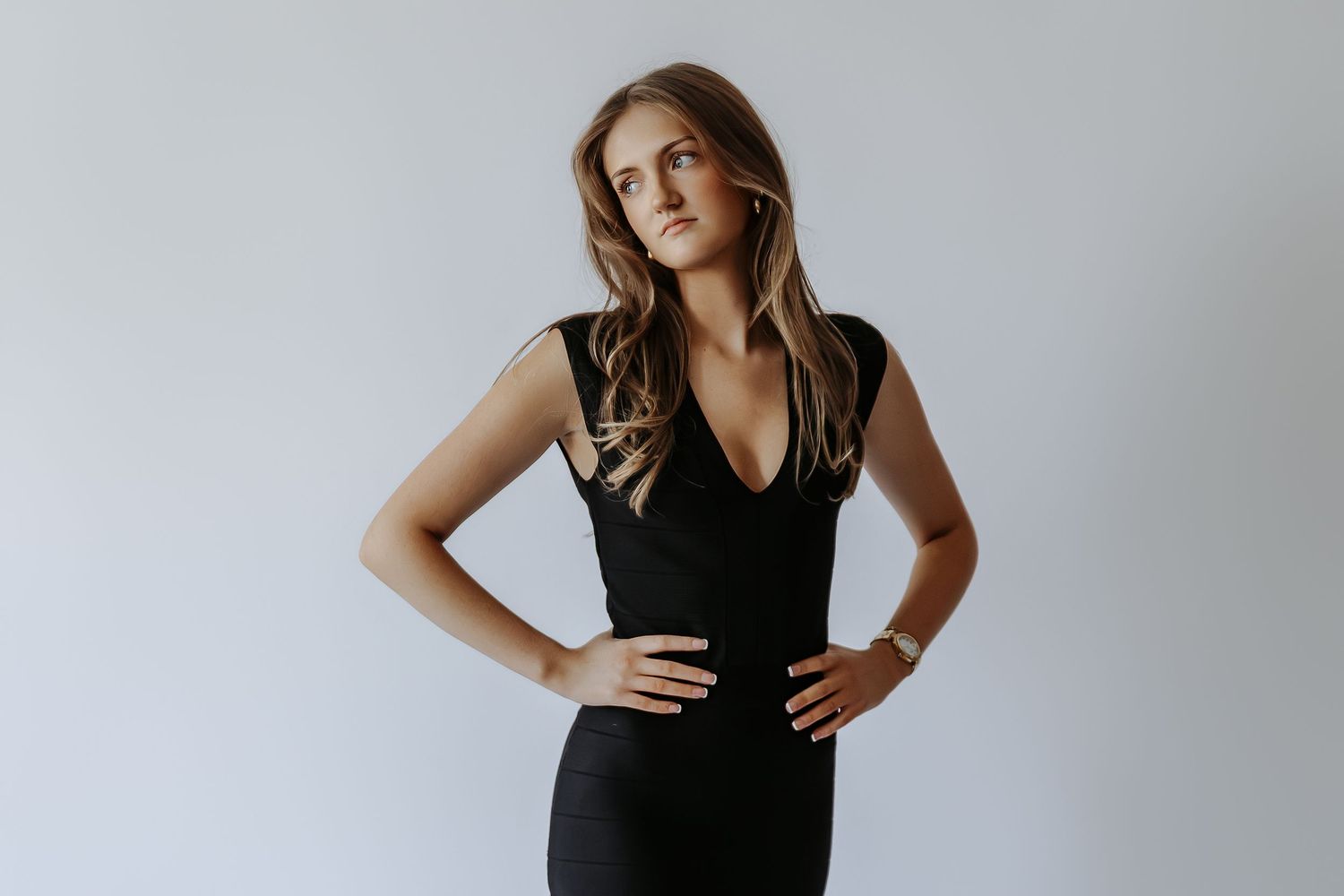 Young woman in a sleek black sleeveless dress posing confidently with hands on hips against a light background.