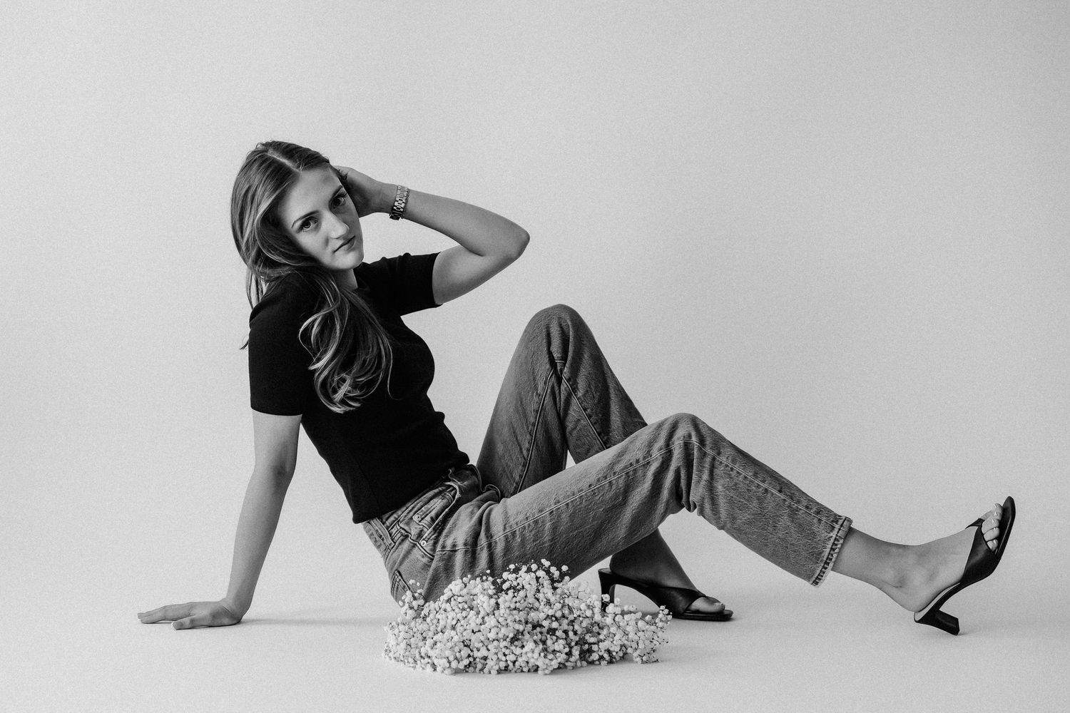 Young woman in black top and jeans reclines on studio floor beside white flowers in black and white photo.