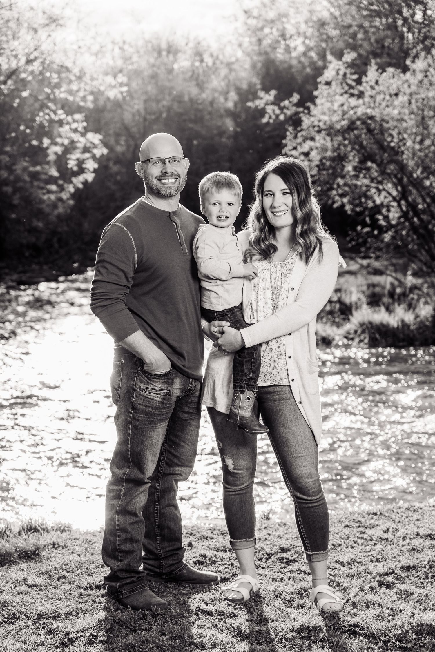 Black and white family portrait of a bald man, woman, and toddler posing outdoors near a sunlit river.