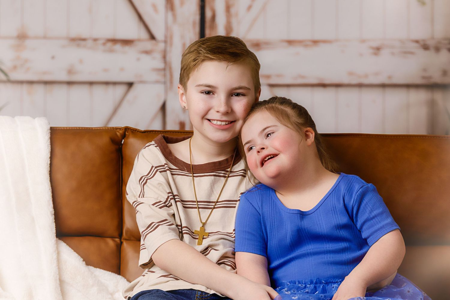 Two children, a boy and a girl with Down syndrome, smile warmly while sitting together on a brown leather couch.