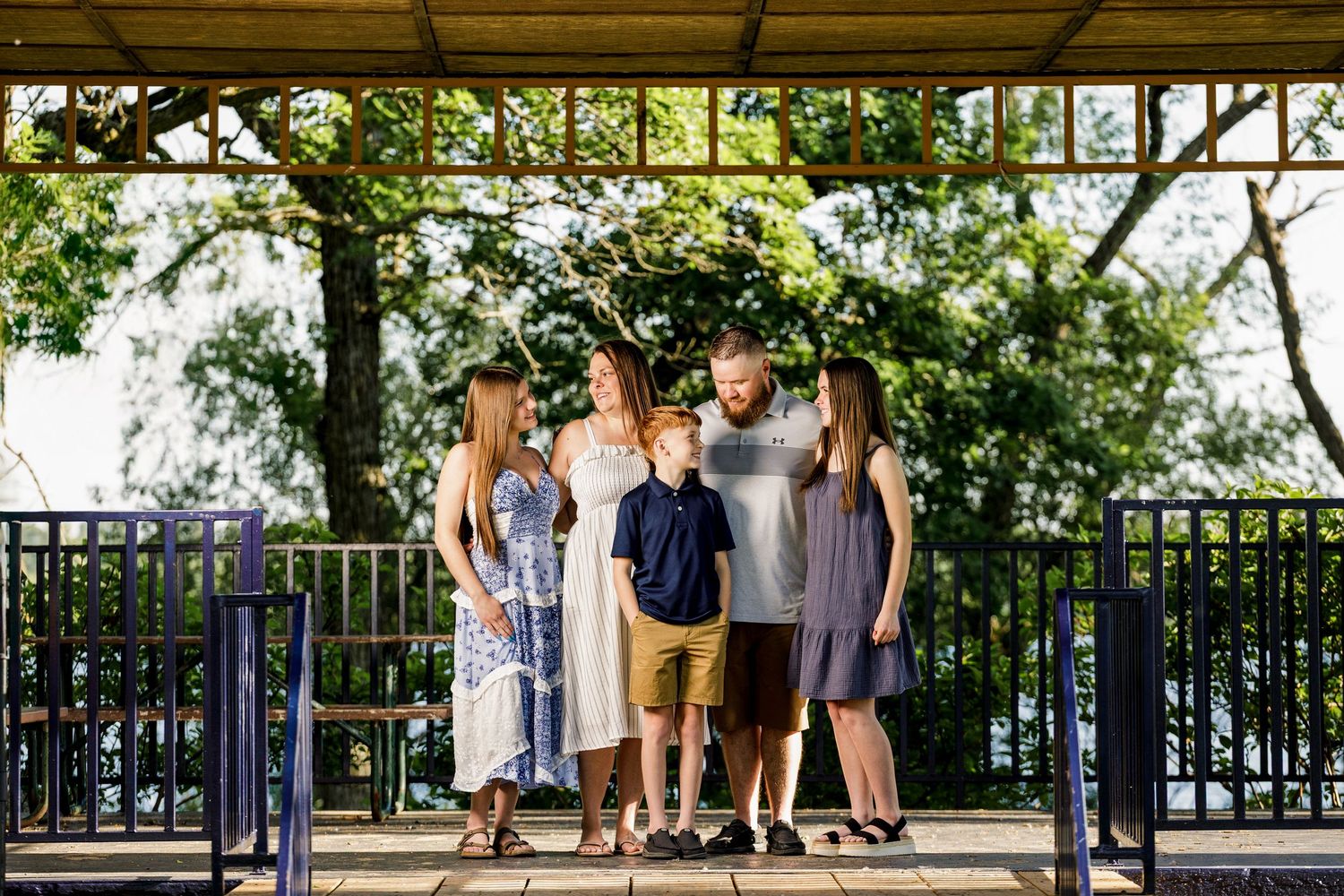 Family of five posing together under a vine-covered pergola outdoors on a sunny day.