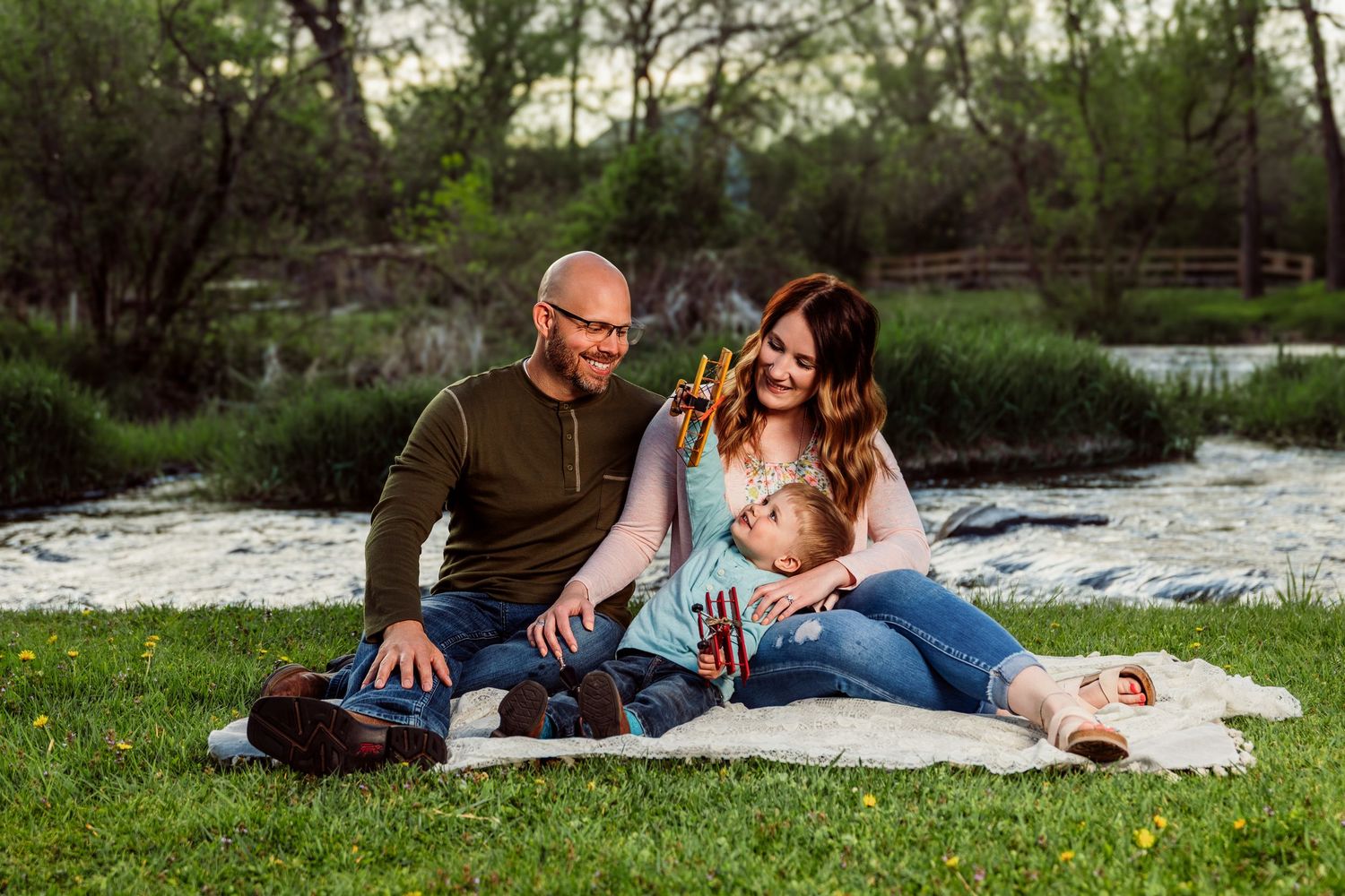Family of three sitting on a blanket by a river in a lush green park during golden hour.