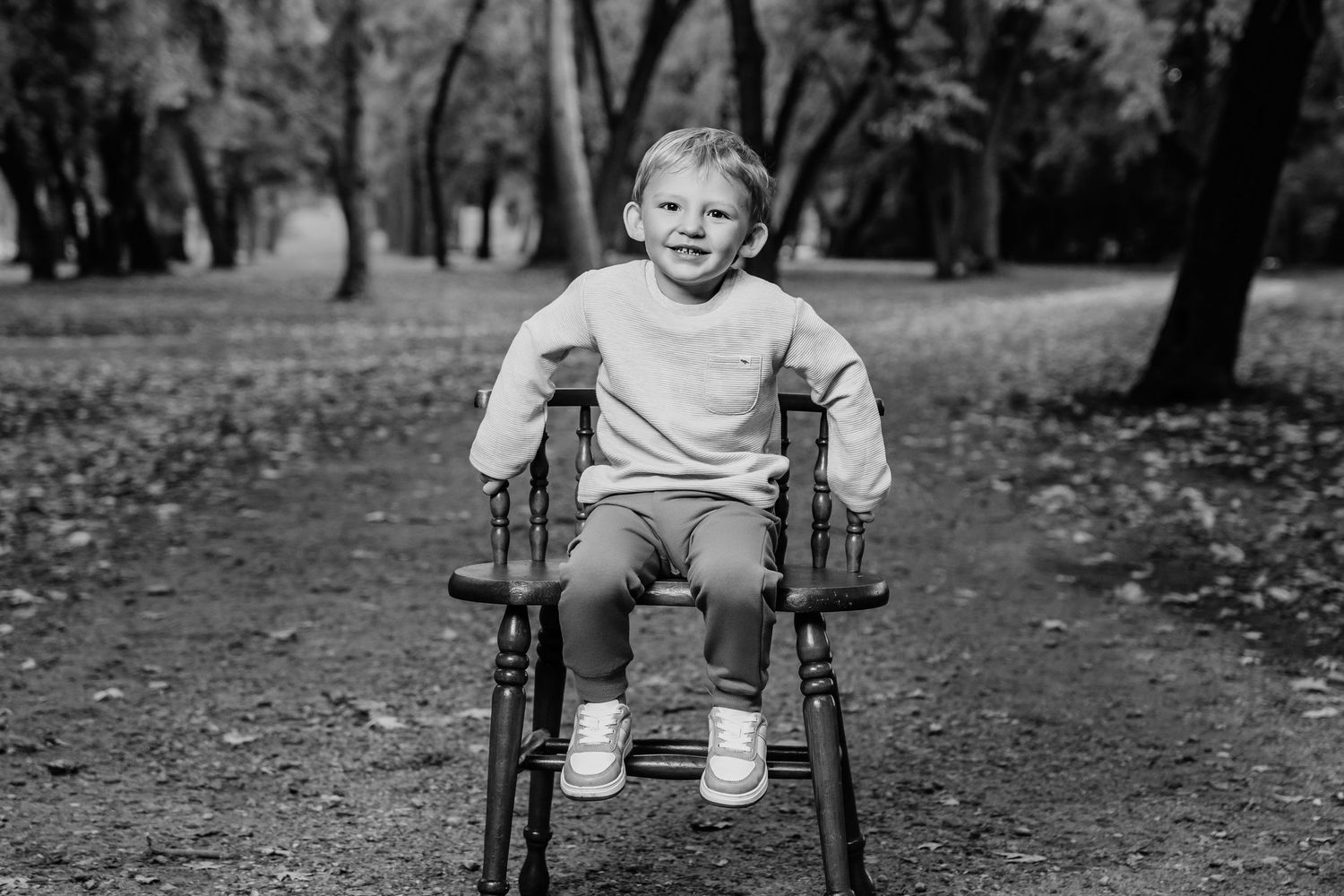 A child sits smiling in an antique wooden chair on a leaf-covered path in the woods.