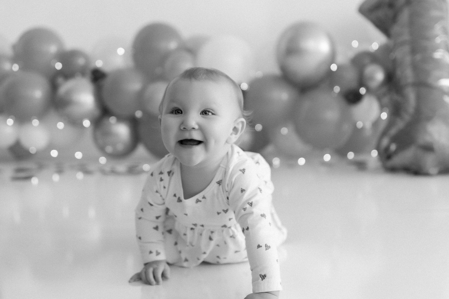 Monochrome capture of baby crawling with festive balloon display behind.