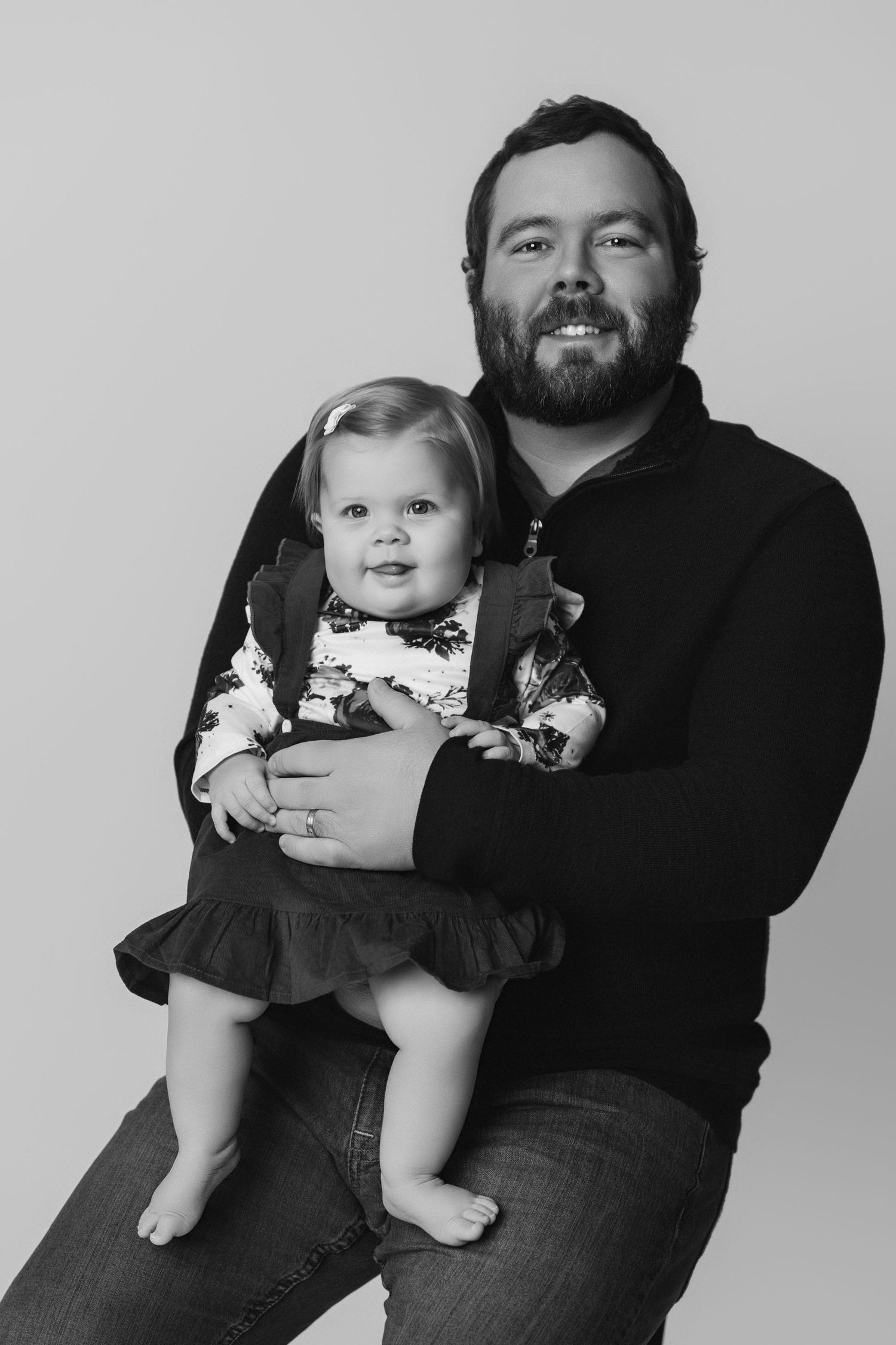 Black and white portrait of a father holding a baby girl wearing a floral dress and bow.