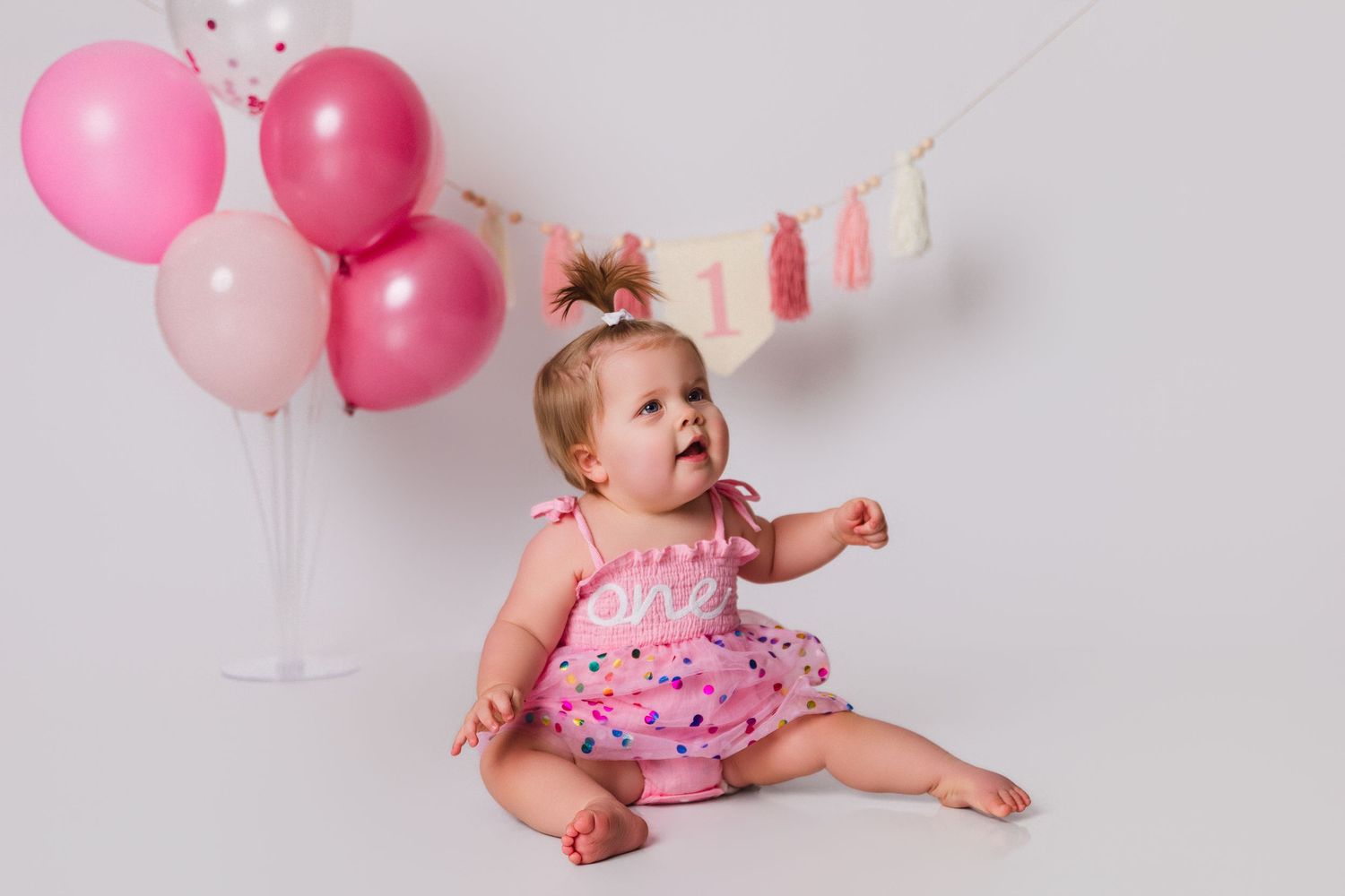 Baby in pink polka dot outfit sitting with pink balloons and party decorations.