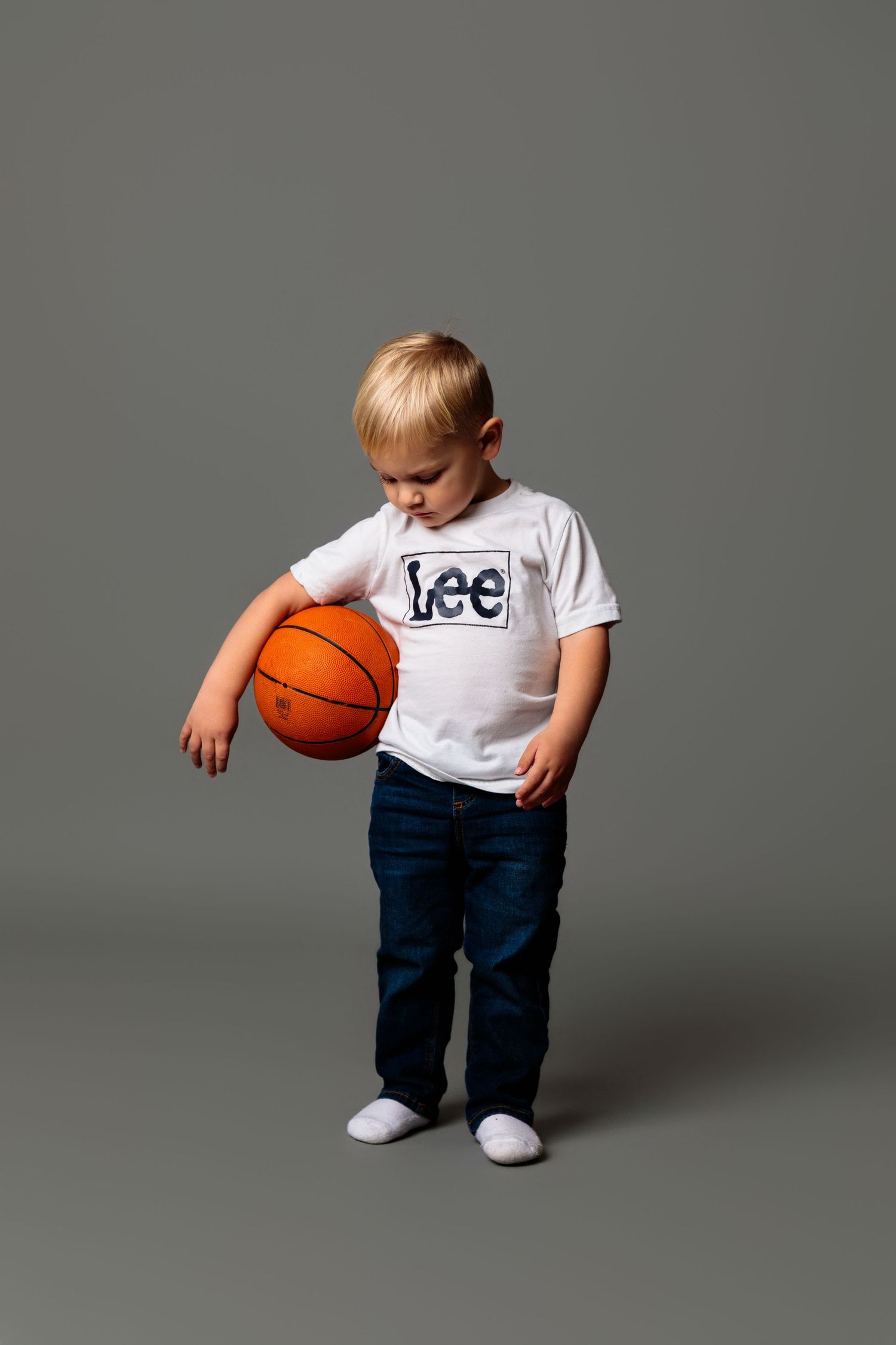 Child in white t-shirt and jeans playing with orange basketball against gray backdrop.