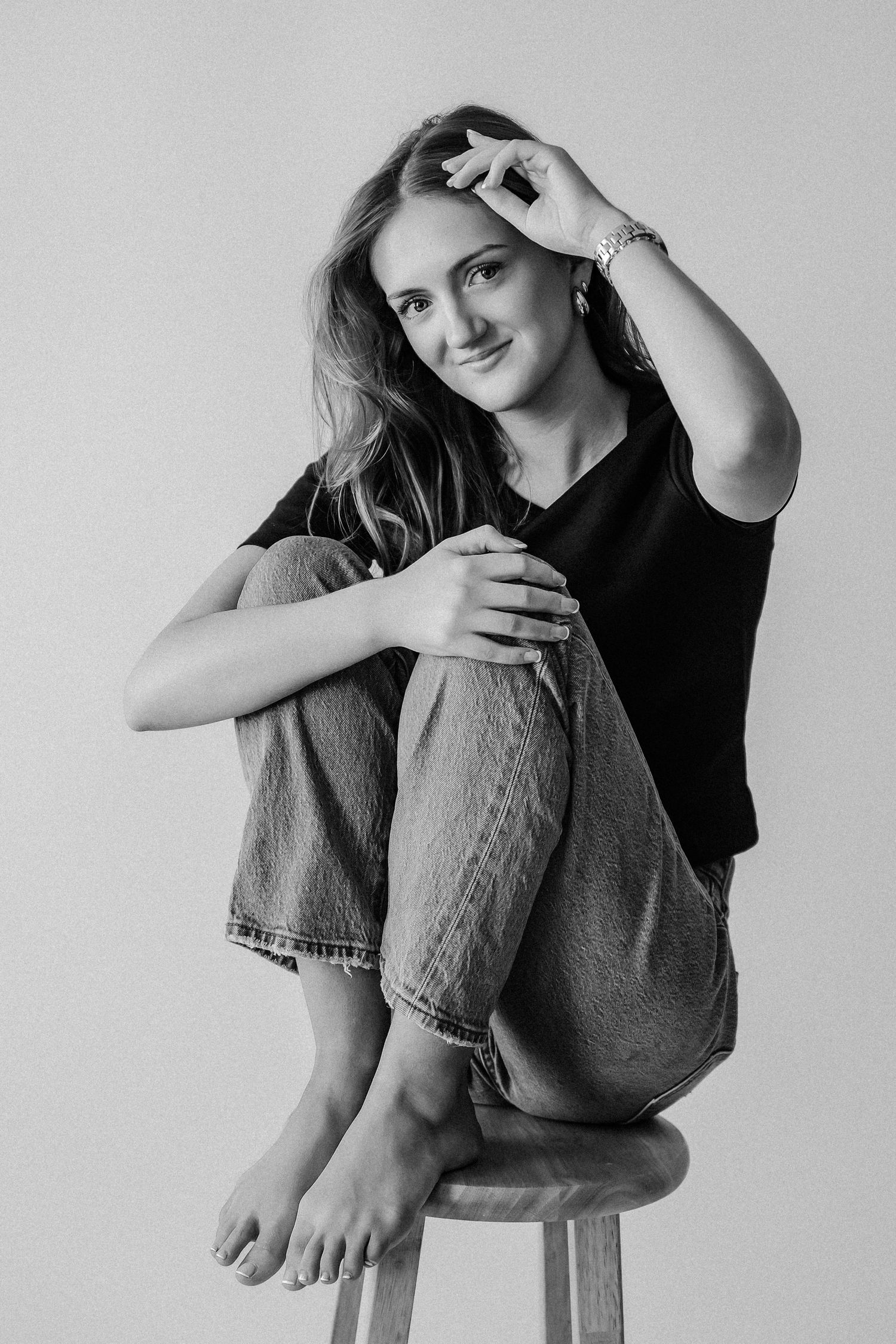 Smiling young woman in black t-shirt and jeans sits relaxed on stool in black and white studio portrait.