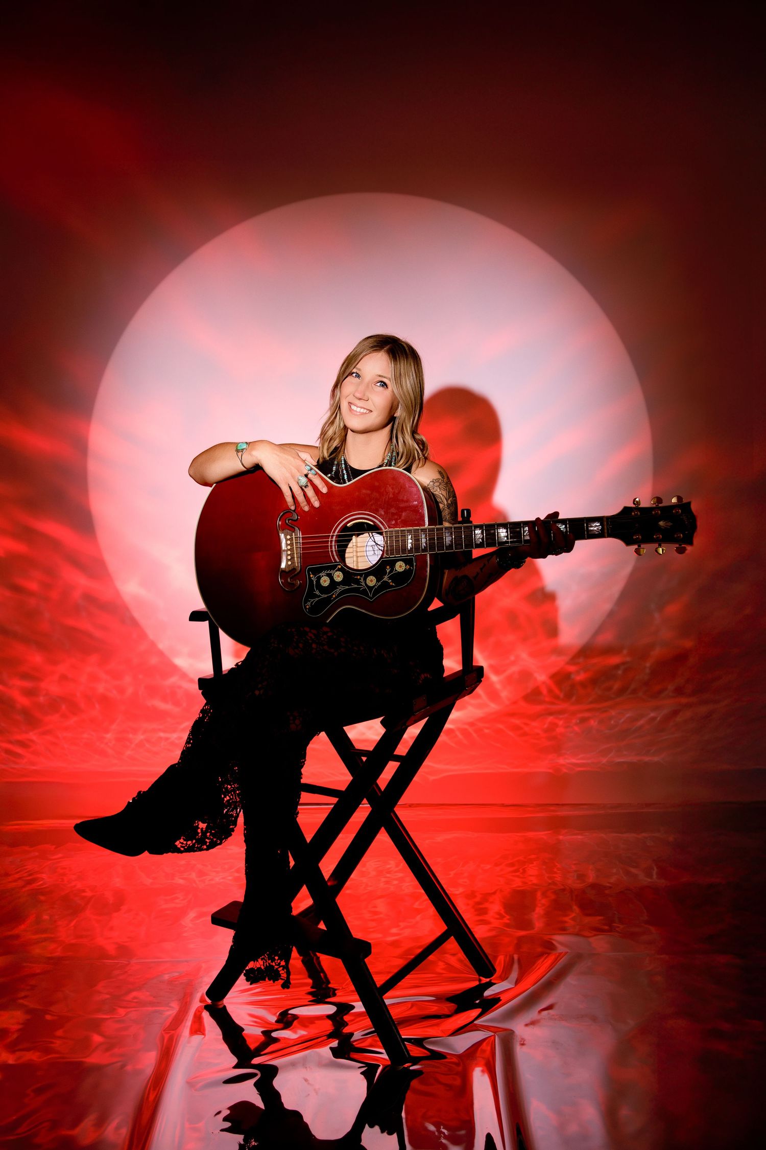 Woman playing red acoustic guitar seated on stool with glowing red moon backdrop and reflection.
