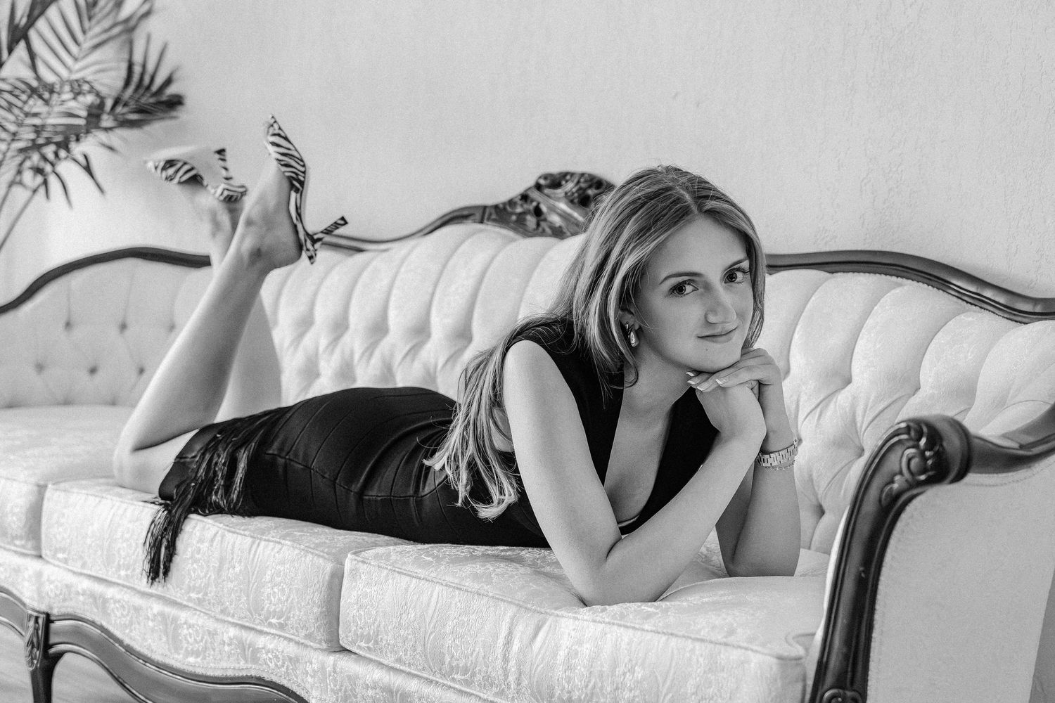 Young woman in black dress reclines gracefully on tufted vintage sofa in black and white photo.