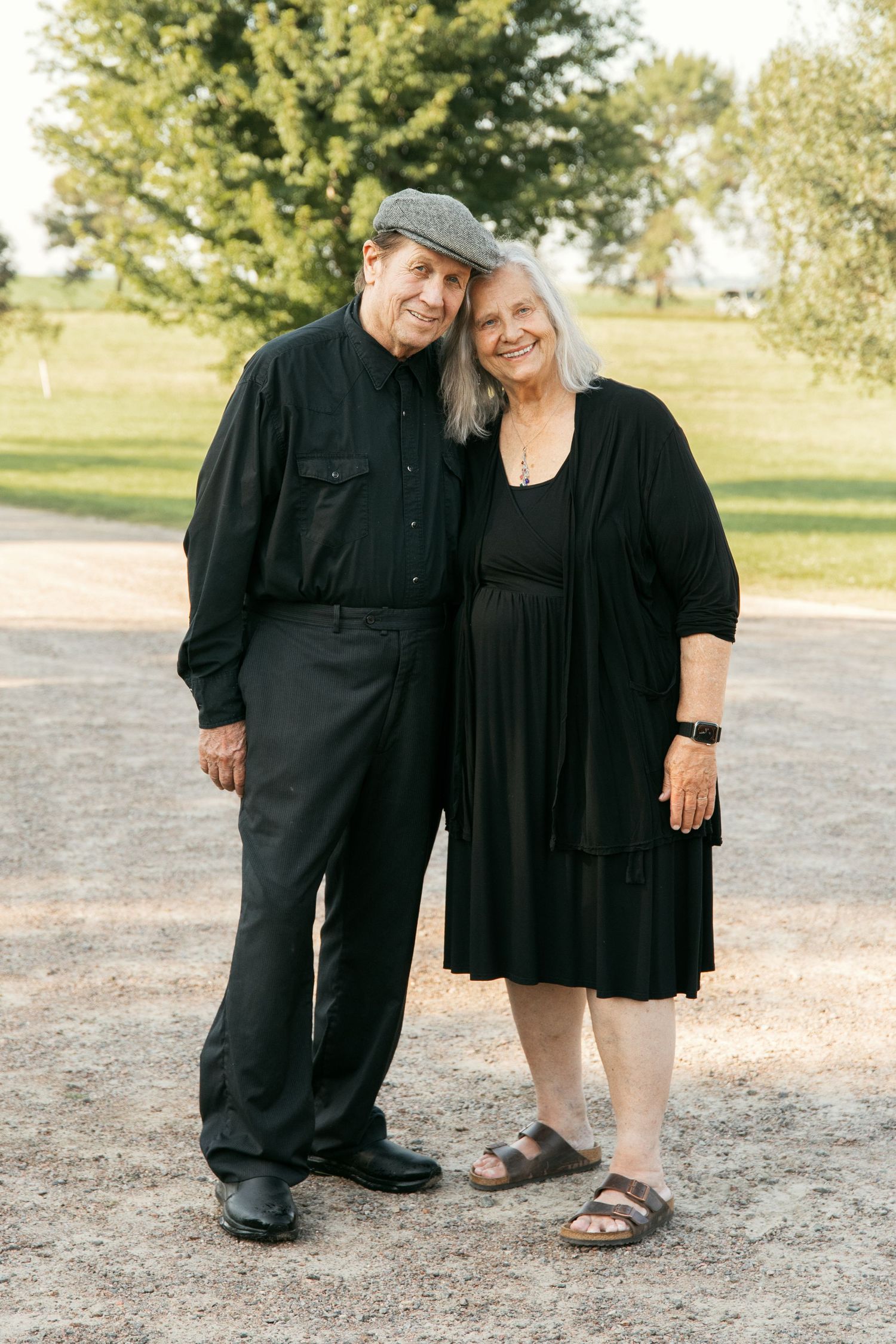 Couple in coordinating black outfits standing together outdoors on a dirt path with trees in background.