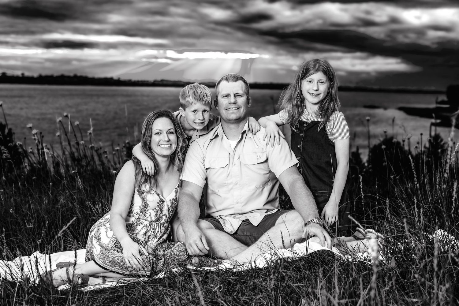 A black and white outdoor family portrait taken on a grassy hillside with dramatic cloudy sky in the background.