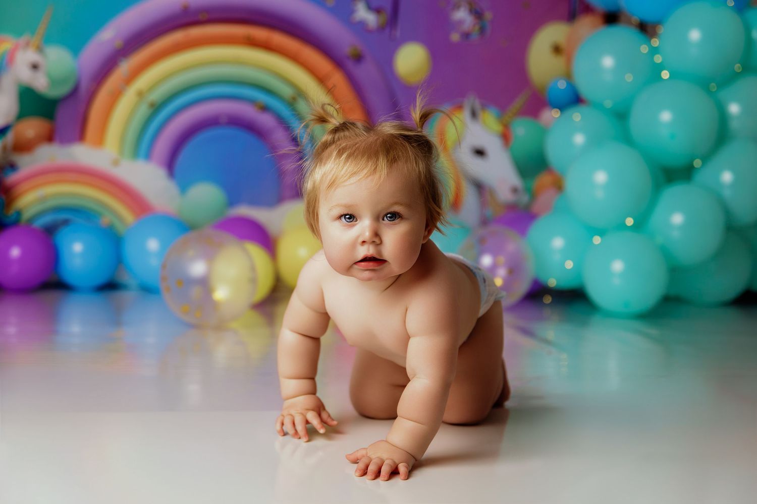 Baby girl in diaper crawls before colorful unicorn rainbow balloon birthday backdrop.