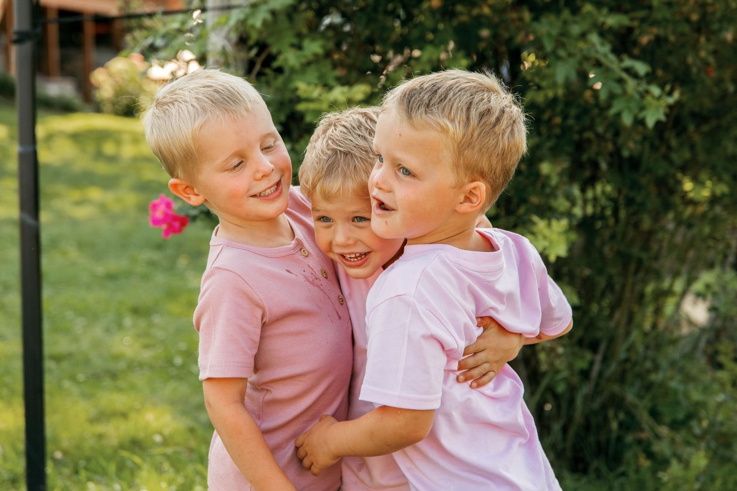 Three children in pink shirts share a joyful embrace while laughing together in a sunny garden setting.