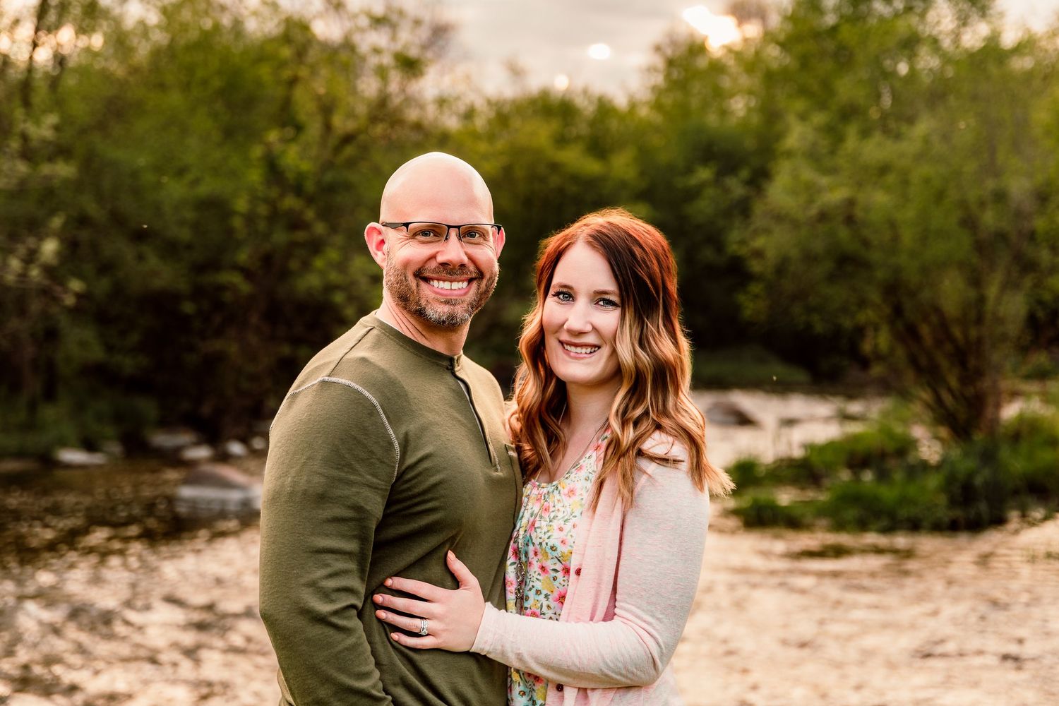 Smiling couple embracing outdoors near a creek with lush green trees in the background during golden hour.