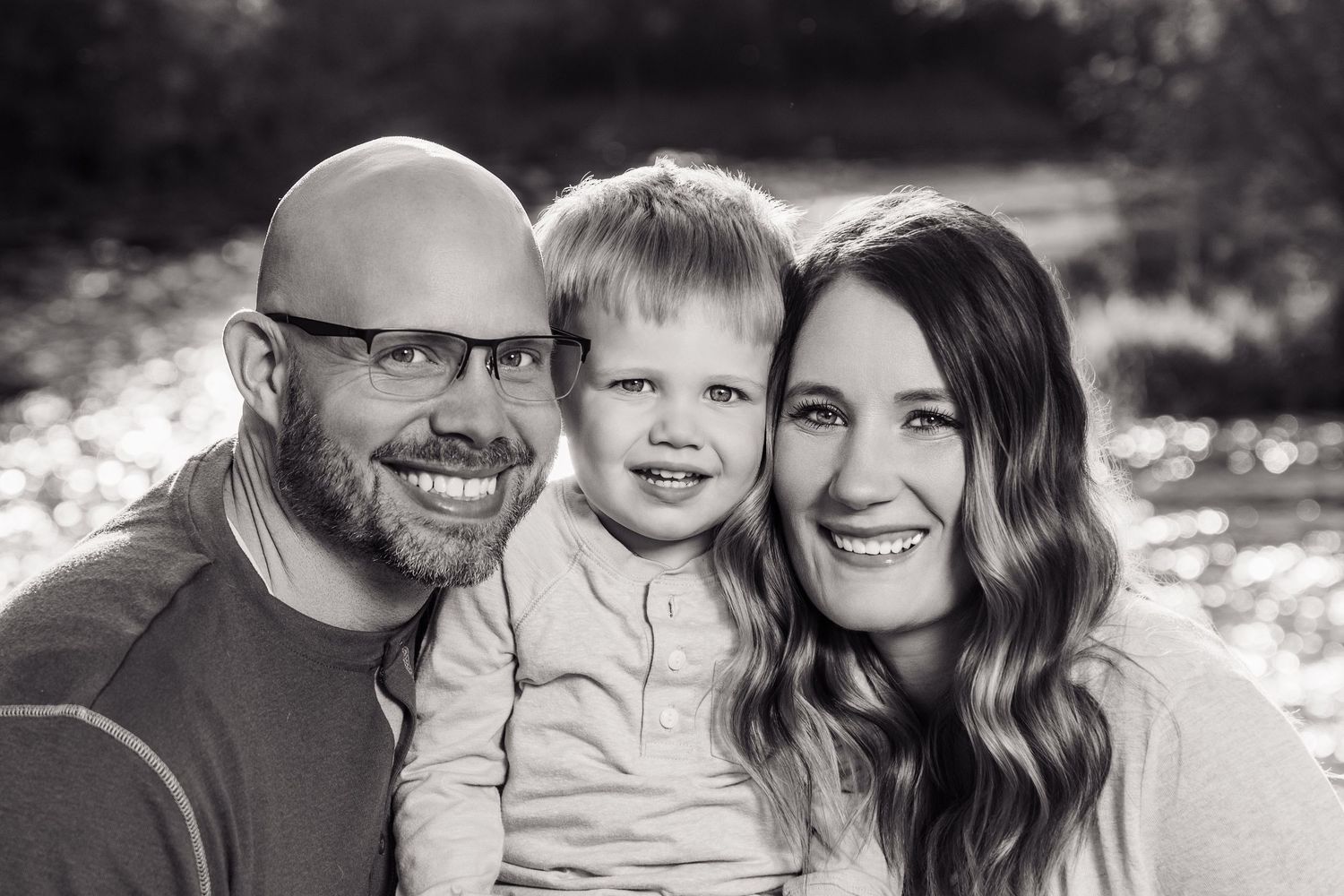 Black and white family portrait of smiling parents with young toddler boy outdoors near a sunlit stream.