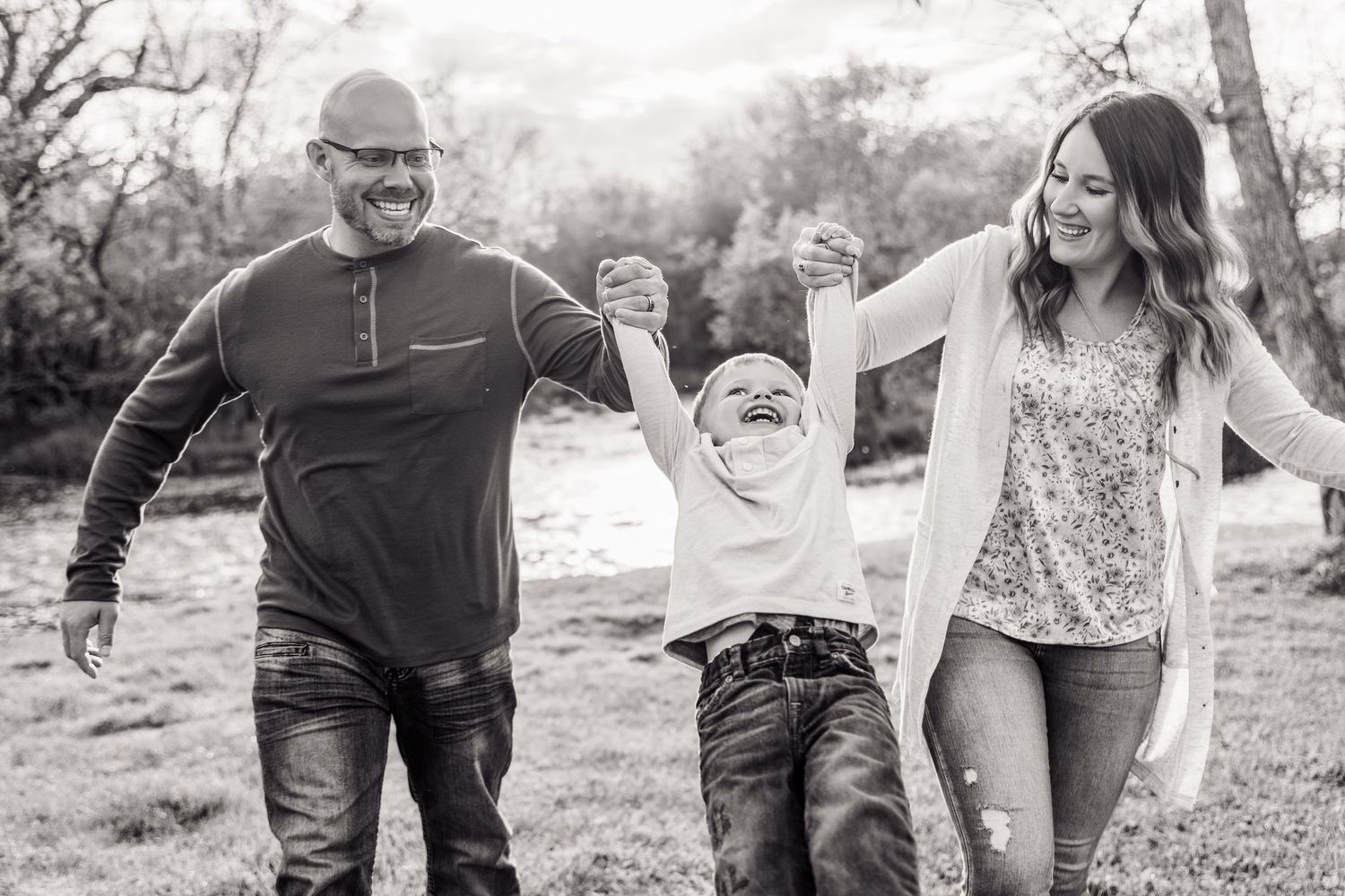 Black and white photo of a joyful family swinging their laughing child outdoors in a park setting.