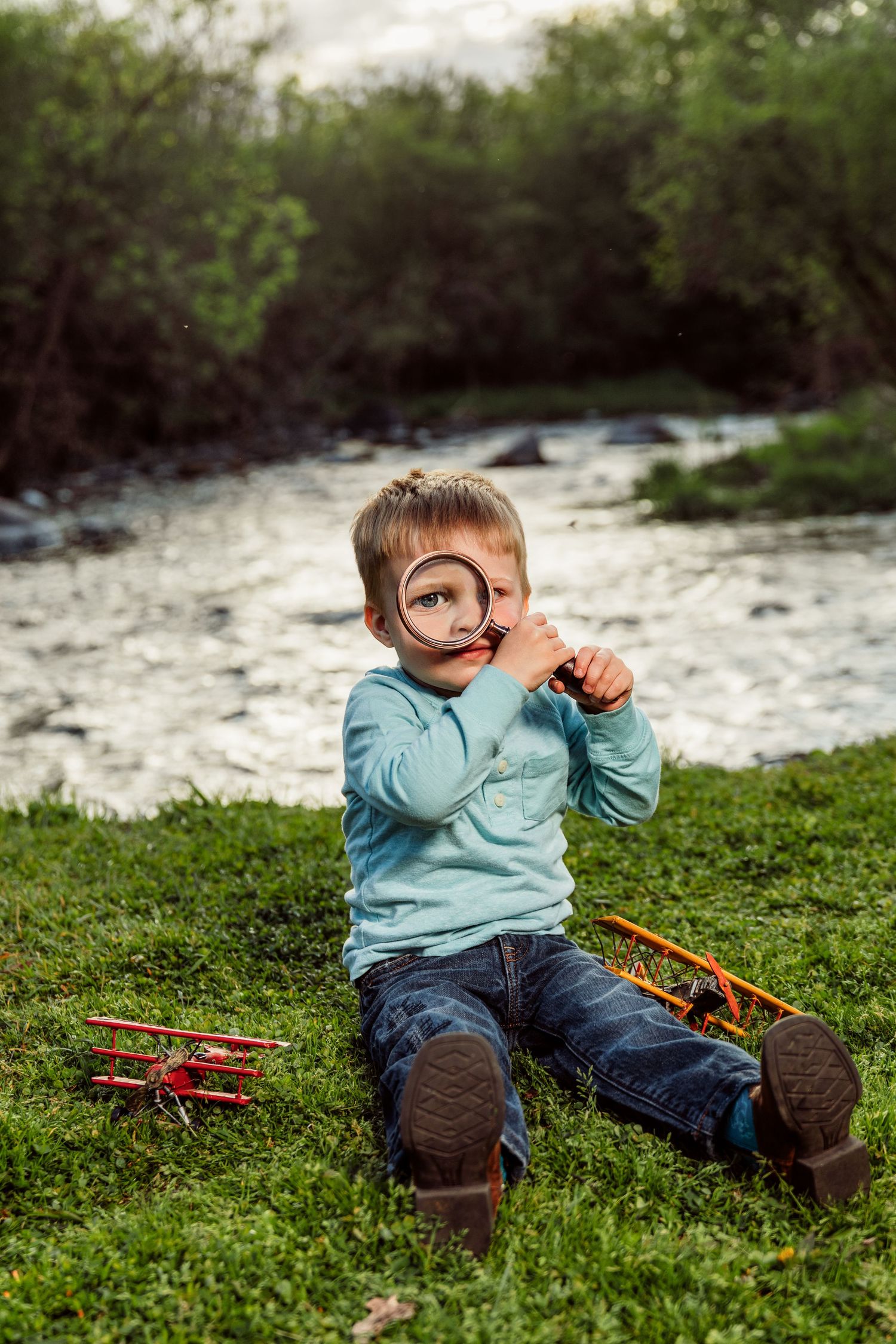 Young boy sitting on grass by a river, holding a magnifying glass to his eye with toy planes nearby.