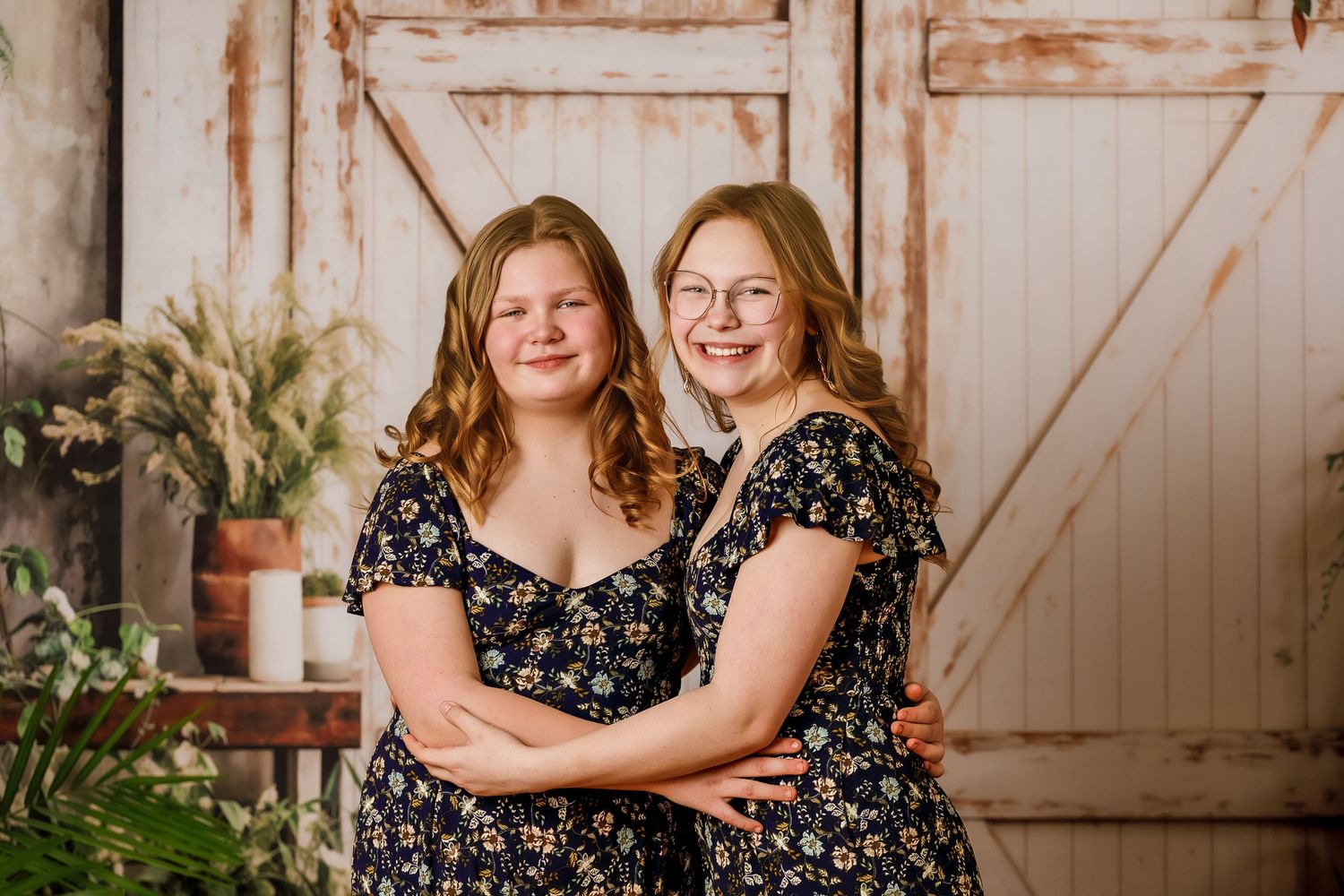 Two young women in matching floral dresses smiling and embracing in front of a rustic barn door backdrop.
