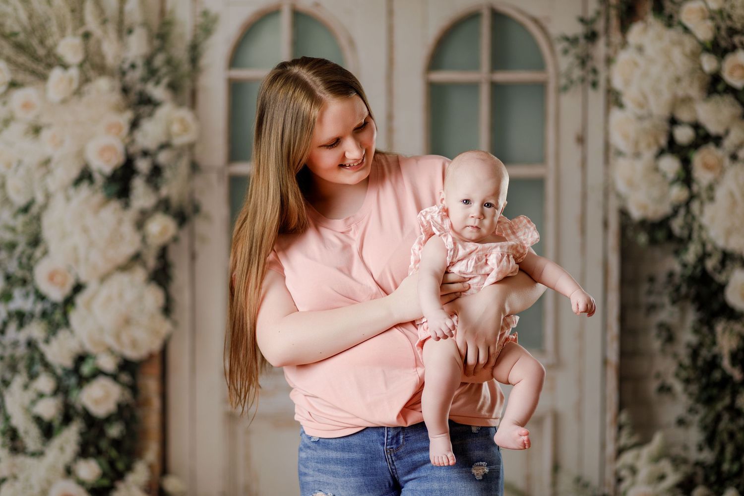A young mother in a pink top holds her baby girl in a floral studio portrait setting.