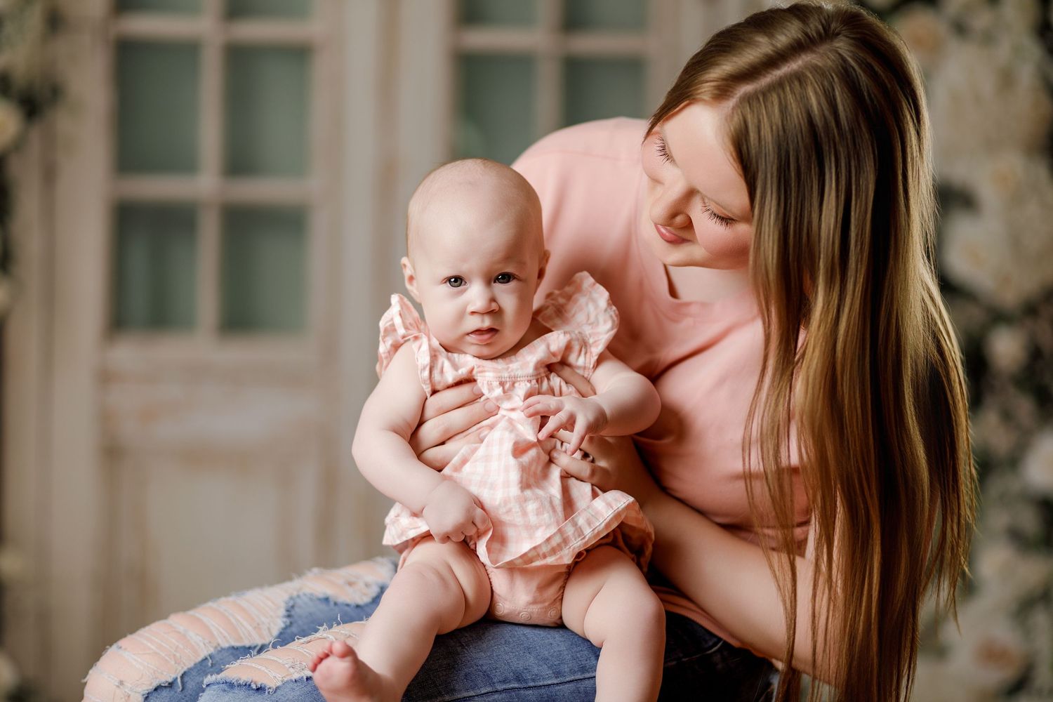 Young mother in pink top lovingly holds her baby girl in a pink floral dress during a studio photo session.