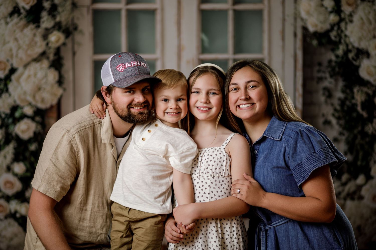 Family of four smiling together in front of a floral backdrop in a professional portrait photo.