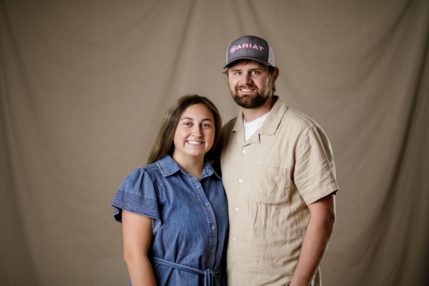 Smiling couple poses together in a casual color portrait against a neutral tan backdrop.