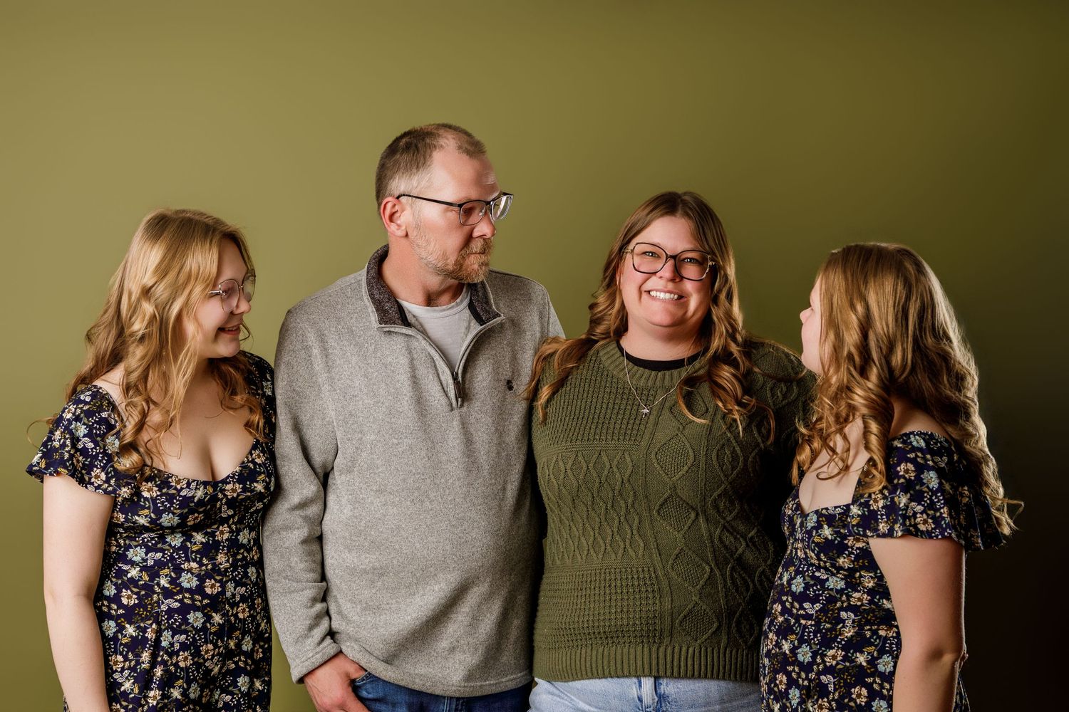 Family of four smiling together against an olive green background in a warm and candid portrait photo.