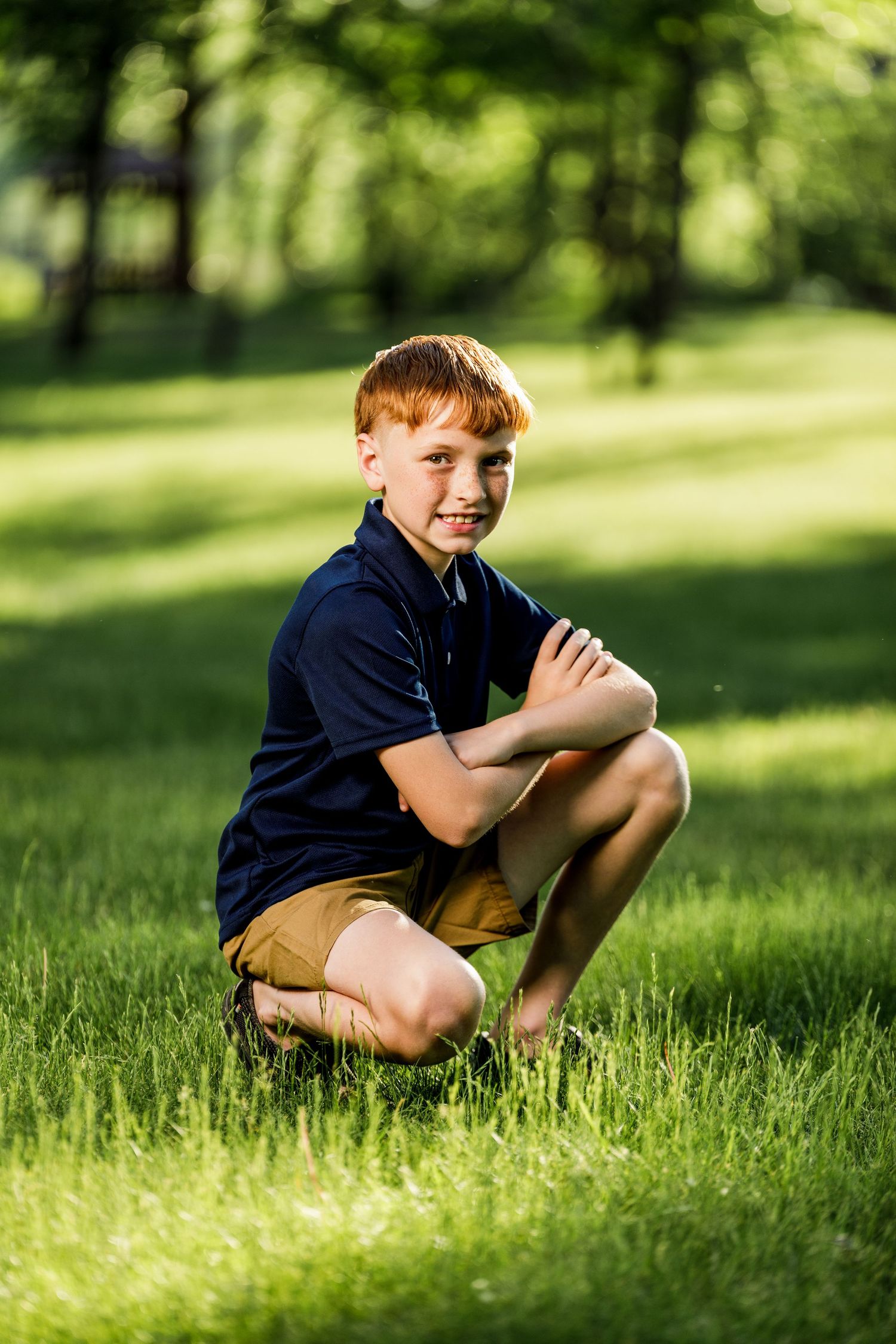 Young redhead boy crouching on green grass in sunny park wearing navy polo and khaki shorts.