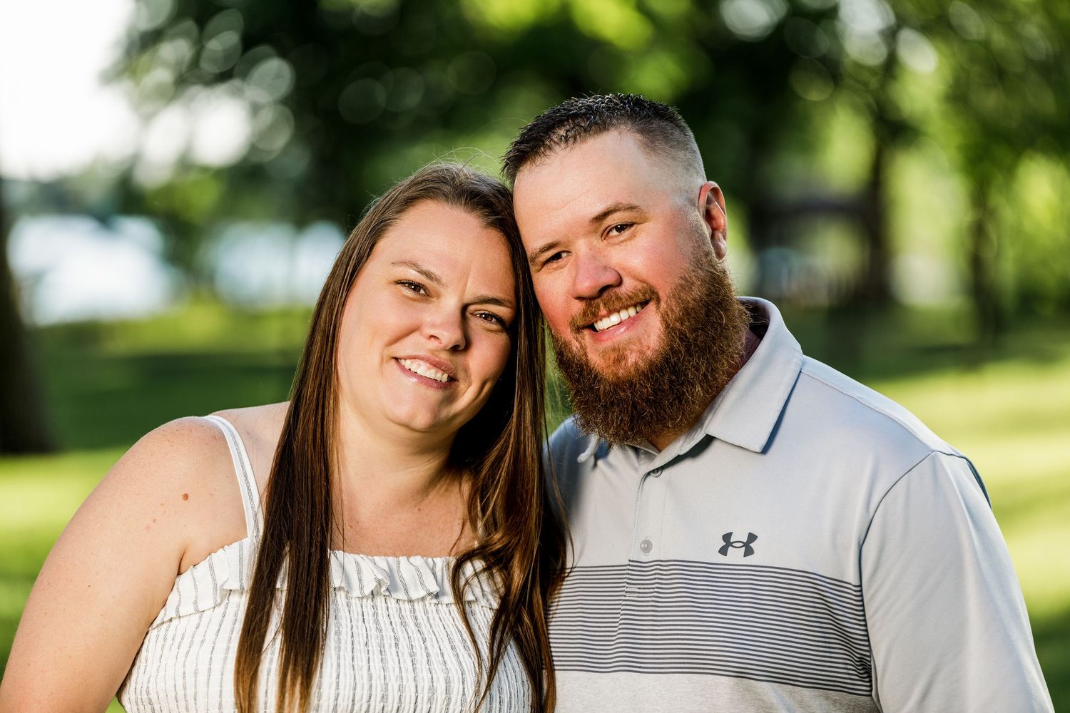 Smiling couple posing together outdoors with a green park and lake in the background.