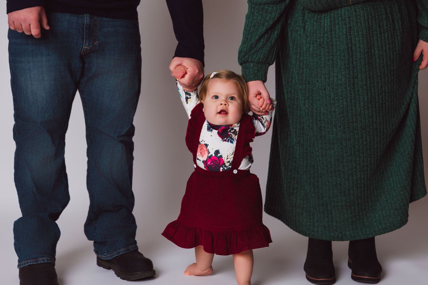 A small child in a burgundy dress holds hands with adults while learning to walk.