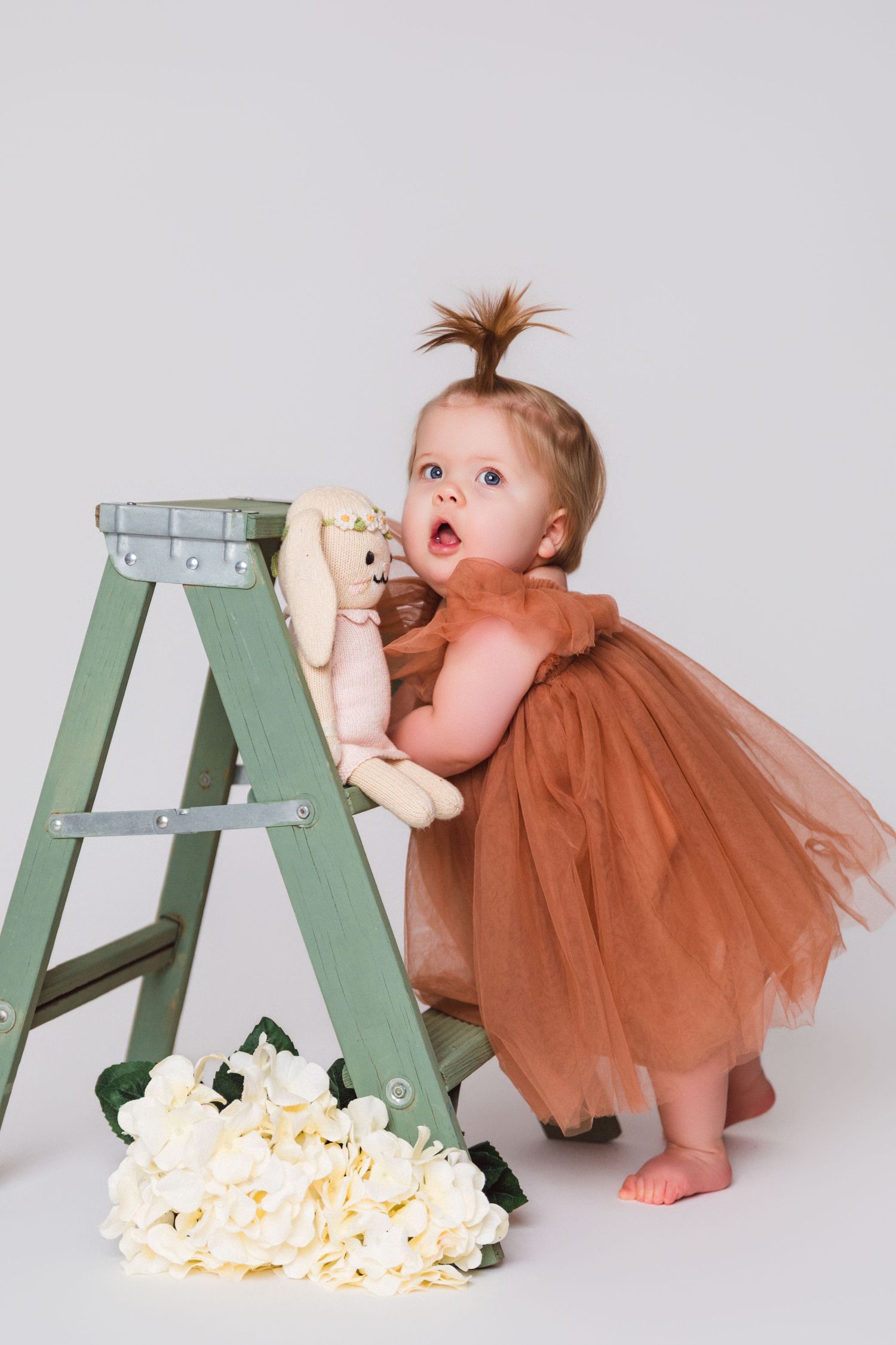 Baby in brown tulle dress playing with a green ladder and scattered flower petals on white background.