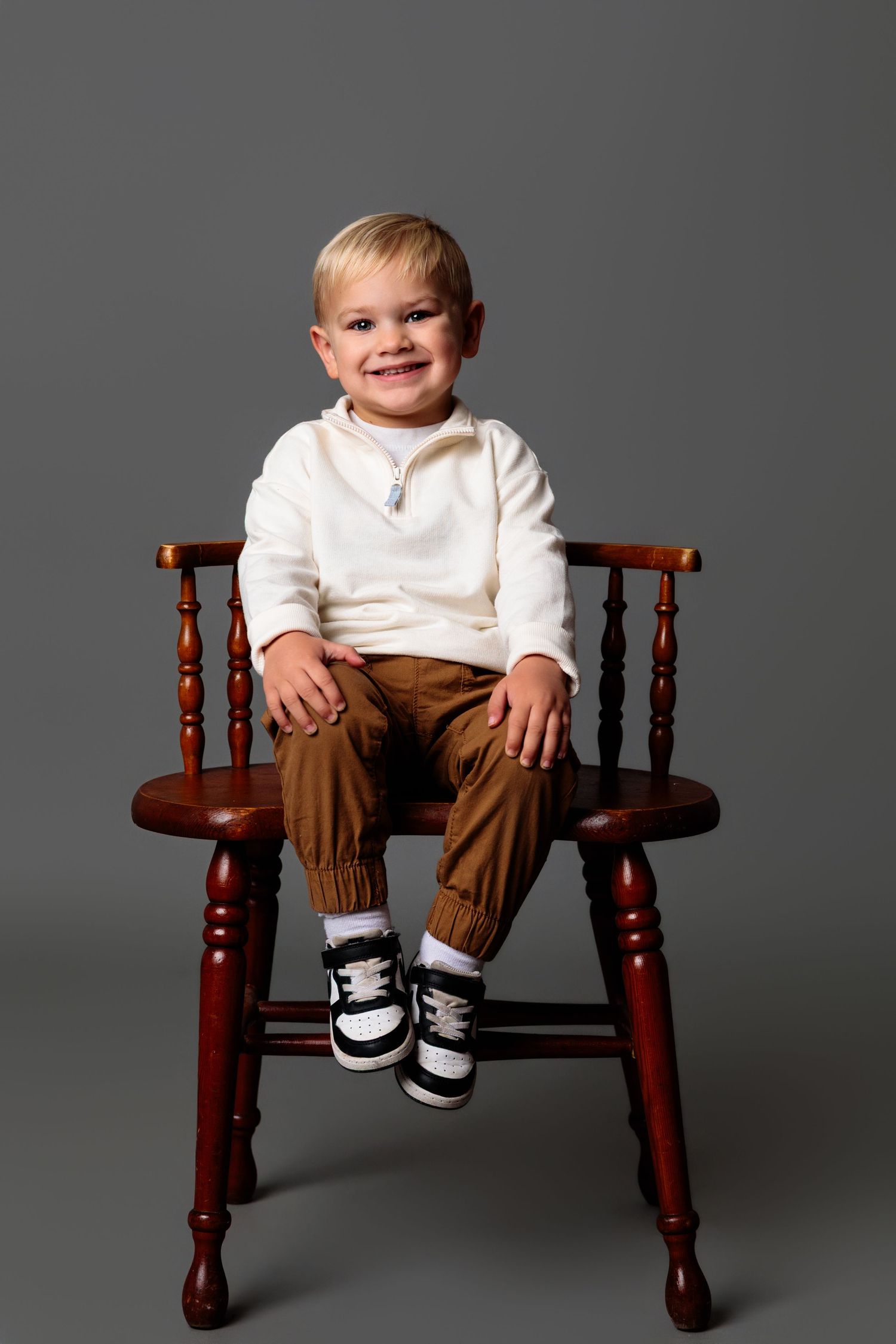 A small child sits on a wooden bench wearing brown pants and a white shirt against a gray background.