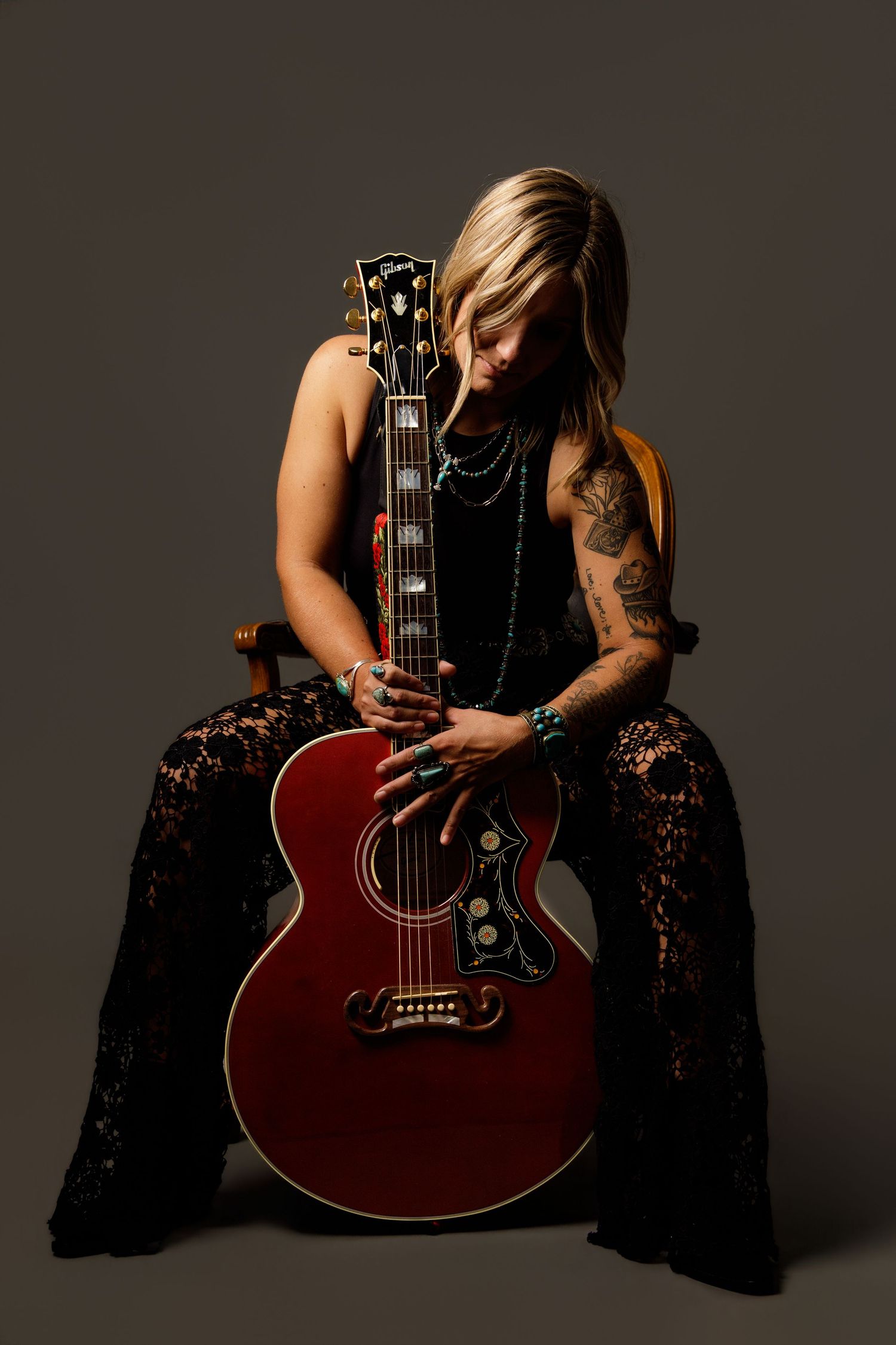 Woman with tattoos in black lace outfit holding red Gibson acoustic guitar while seated in moody studio lighting.
