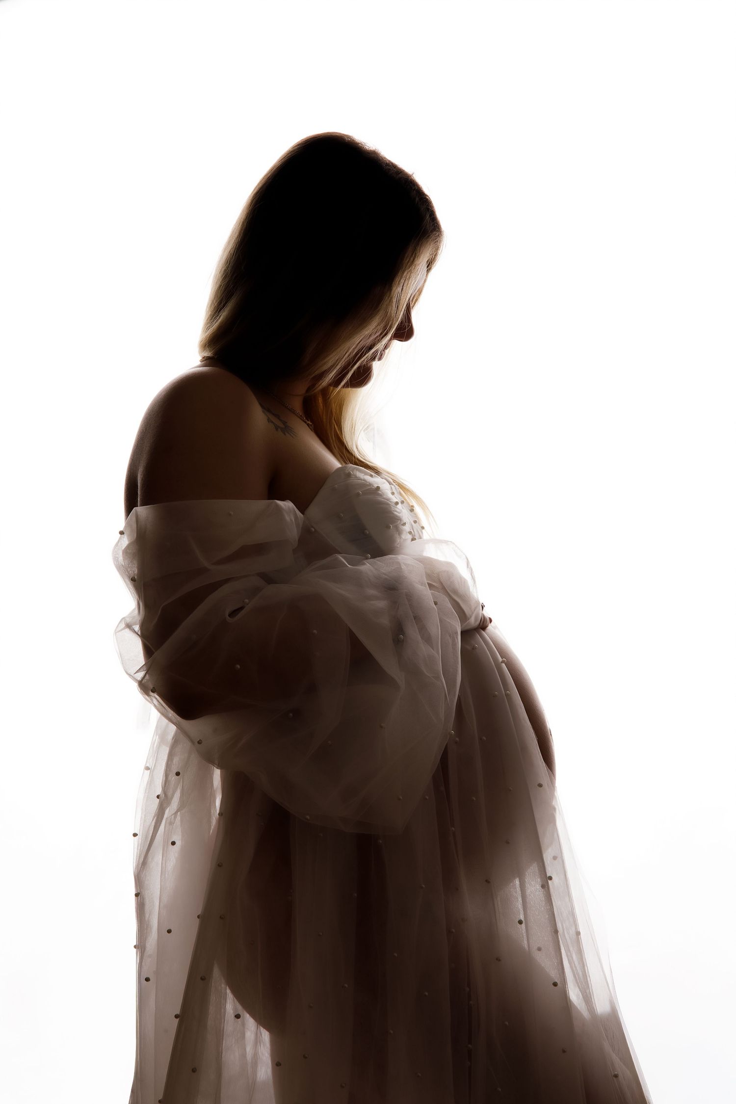 Silhouette of a pregnant woman in a flowing white tulle gown, cradling her baby bump against a bright white background.