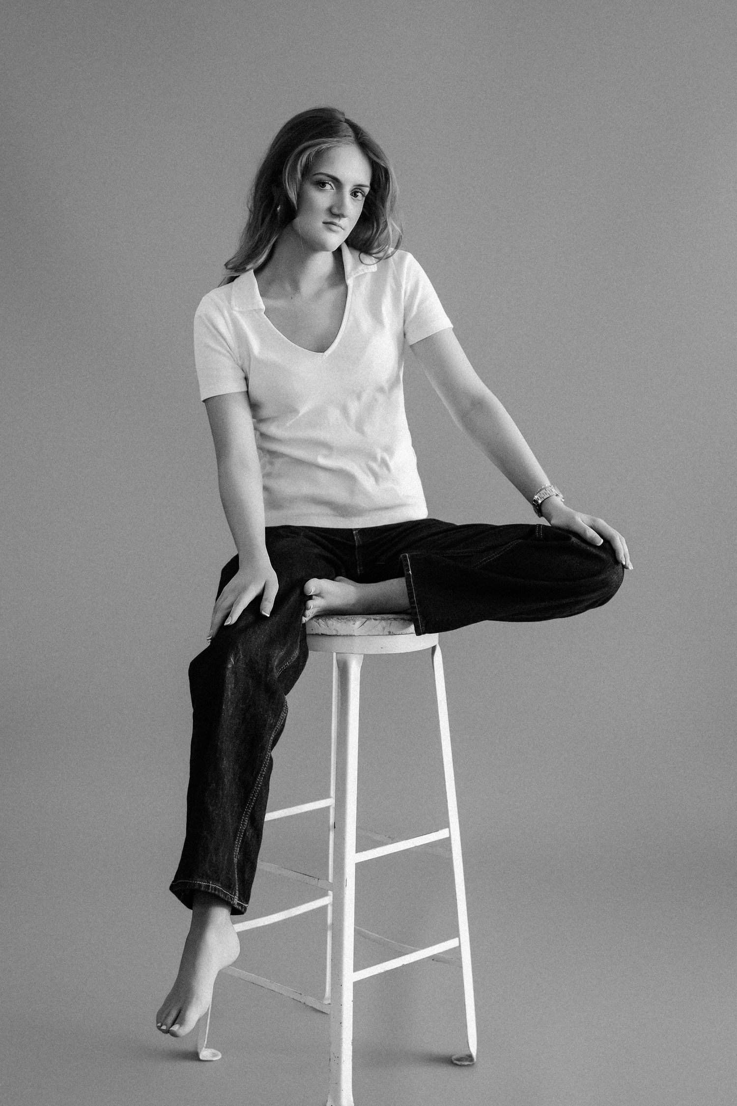 Young woman sitting on white stool barefoot in white t-shirt and dark jeans, black and white.