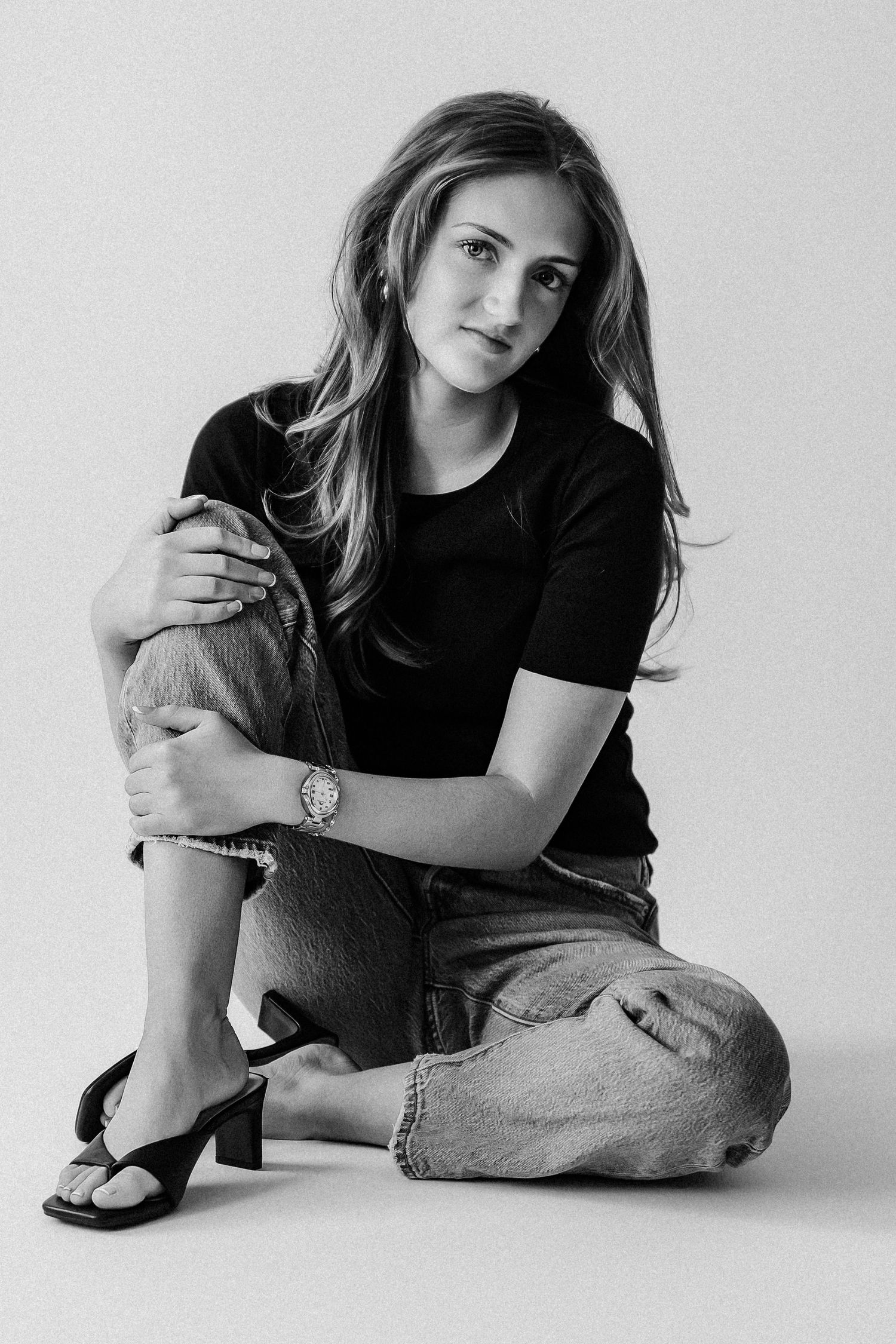 Young woman in black t-shirt and denim jeans sits on floor in classic black and white studio photo.